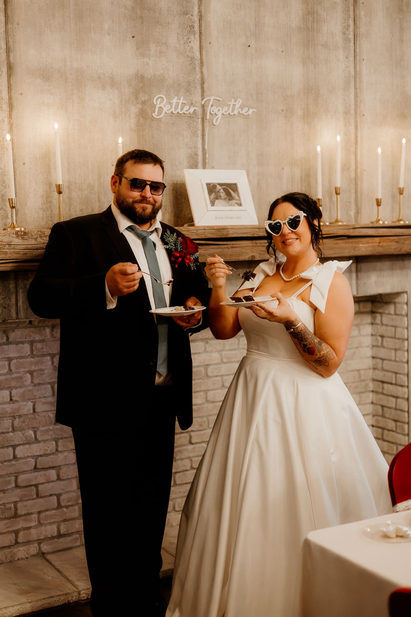 Bride in heart-shaped sunglasses and groom in dark suit and sunglasses eat wedding cake in front of a candlelit mantel and brick fireplace.
