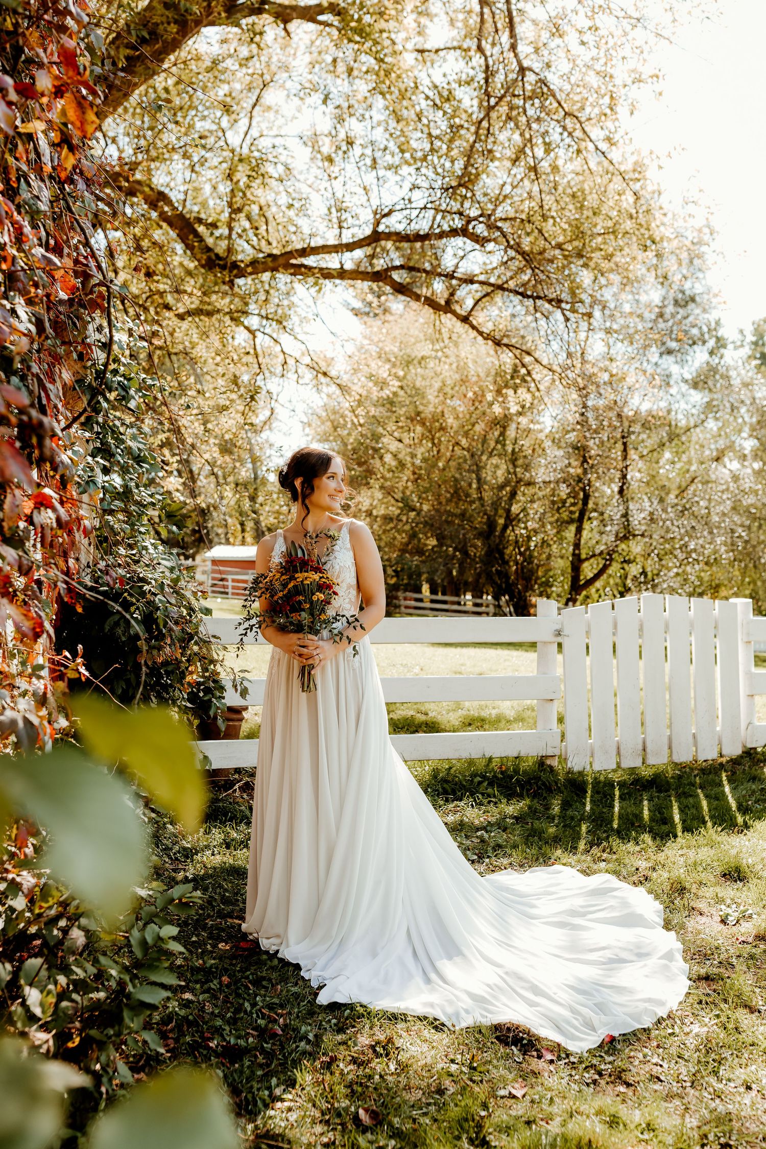 Bride in a flowing white gown holding a colorful bouquet stands on a sunlit lawn beside a white fence and autumn trees.