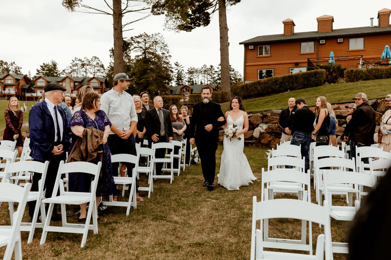 Bride and groom walk hand in hand down the grass aisle past white chairs and smiling guests at an outdoor wedding ceremony.