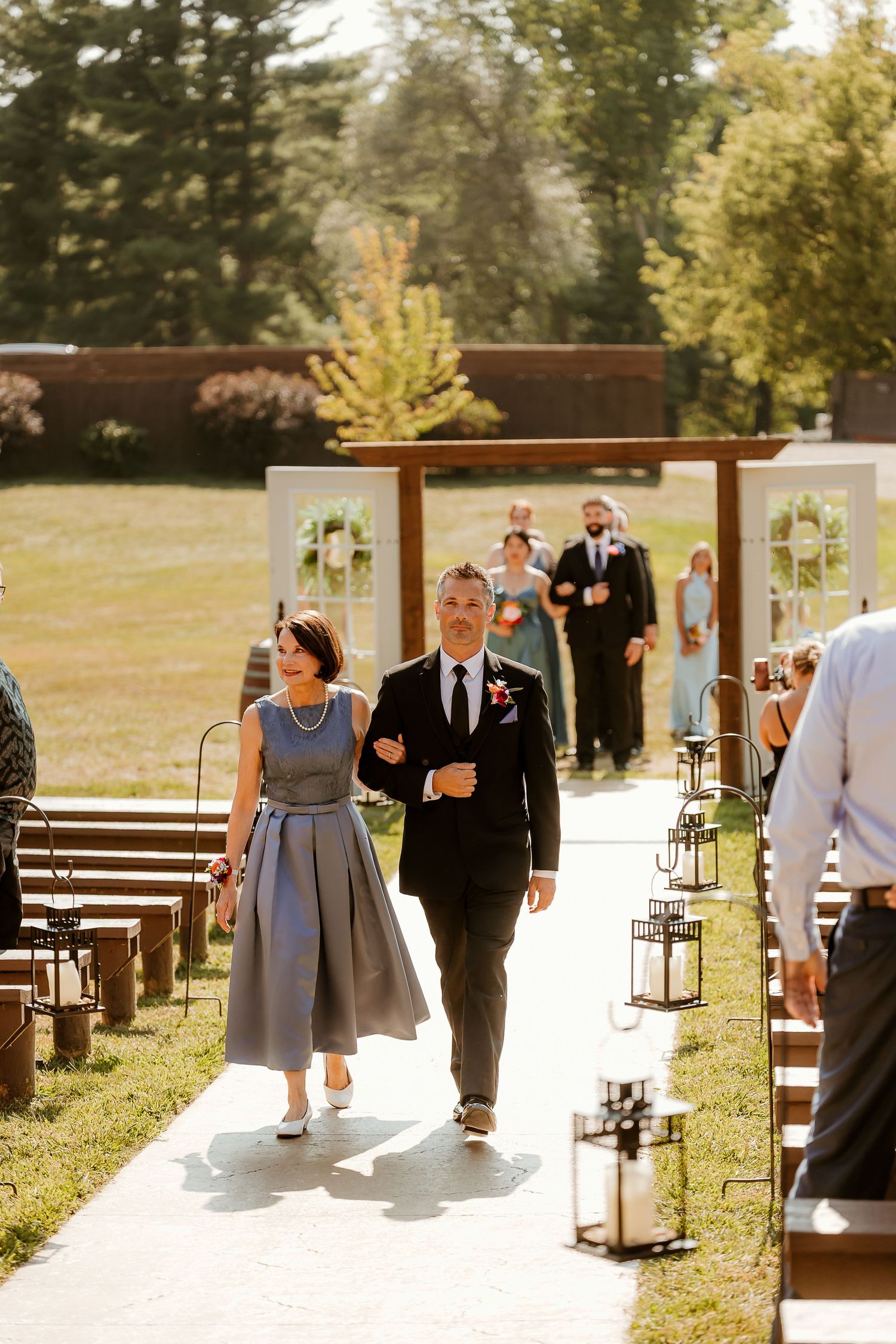 Couple walks down outdoor wedding aisle lined with lanterns and wooden benches, with arch and bridal party visible behind them.