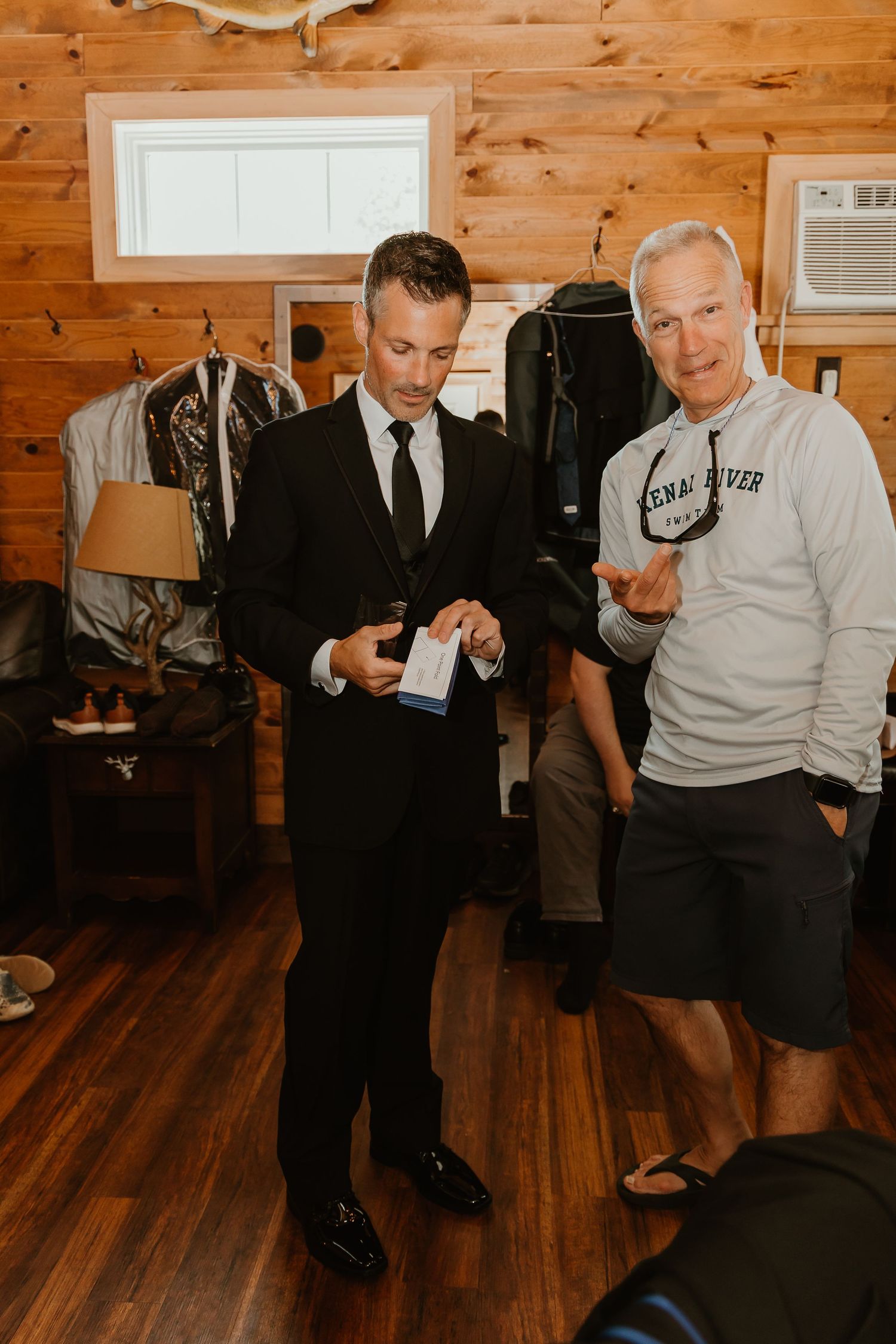 Two men in a wood-paneled room, one in a black suit adjusting his outfit, the other in casual wear gesturing nearby.
