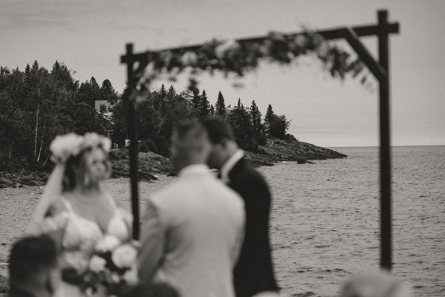 Black and white photo of a couple exchanging vows under a floral arch by the ocean with evergreen trees in the background.