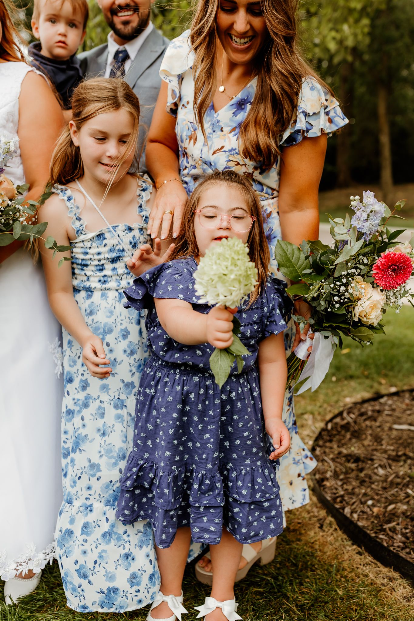 Young flower girls in blue floral dresses hold bouquets at a wedding, smiling alongside the bridal party outdoors.