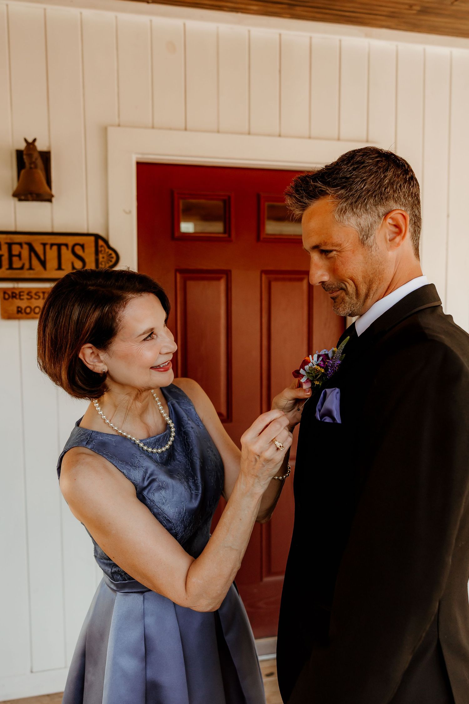 A woman in blue dress pins a boutonniere on a groom in a black tuxedo outside a red door.