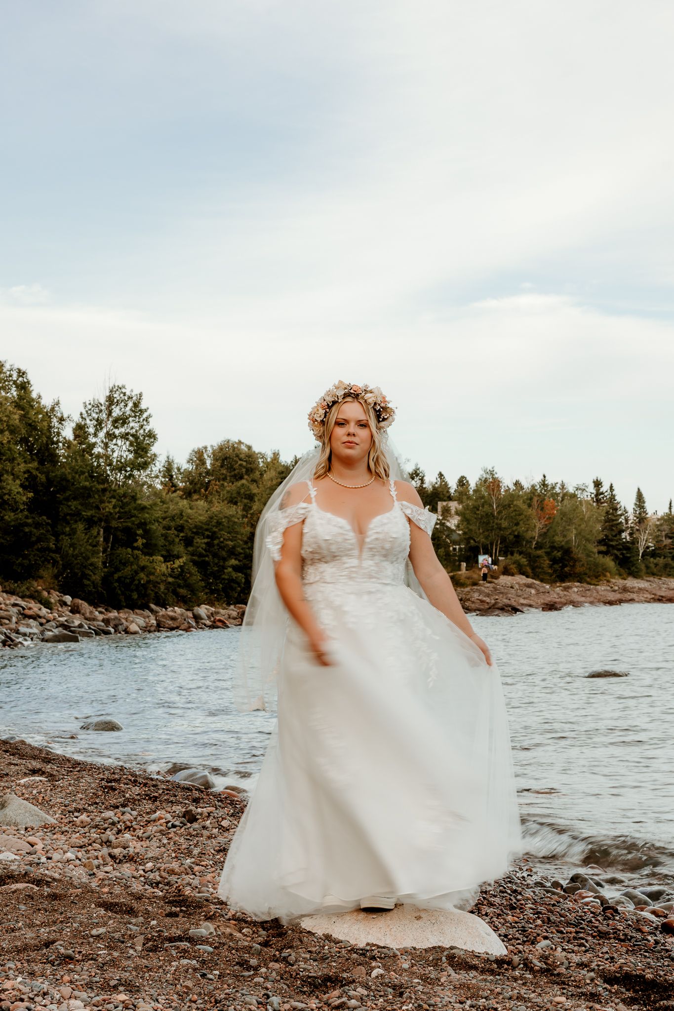 Bride in white lace gown and floral crown poses by rocky lakeside with trees in background.