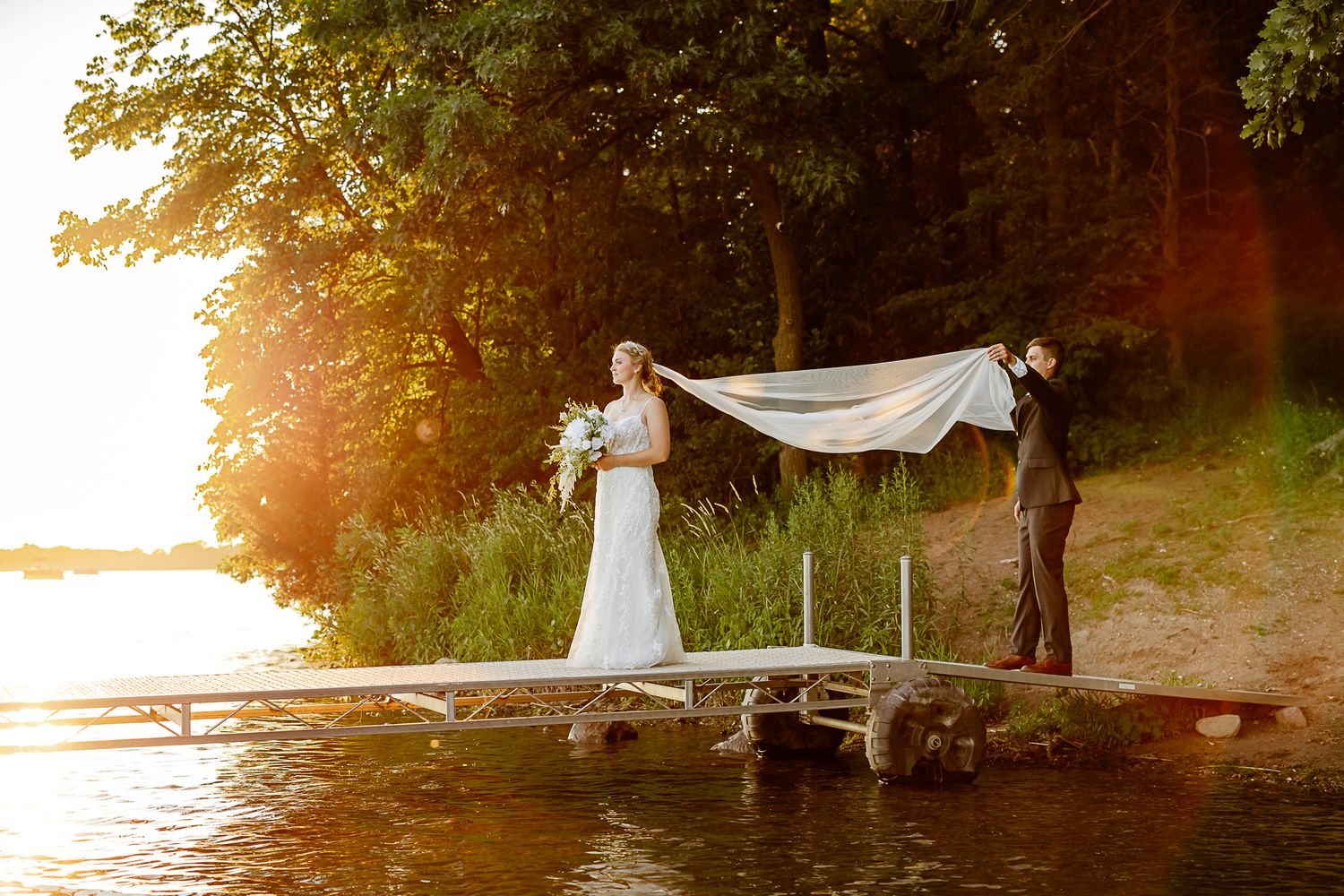 Bride and groom on a lakeside dock at sunset, with veil flowing in the breeze amid lush green trees.