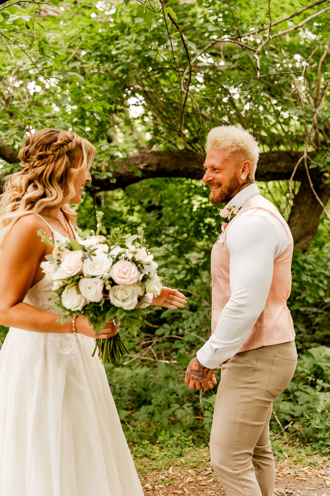 Groom smiling joyfully at bride holding white and pink rose bouquet during outdoor wedding first look ceremony.