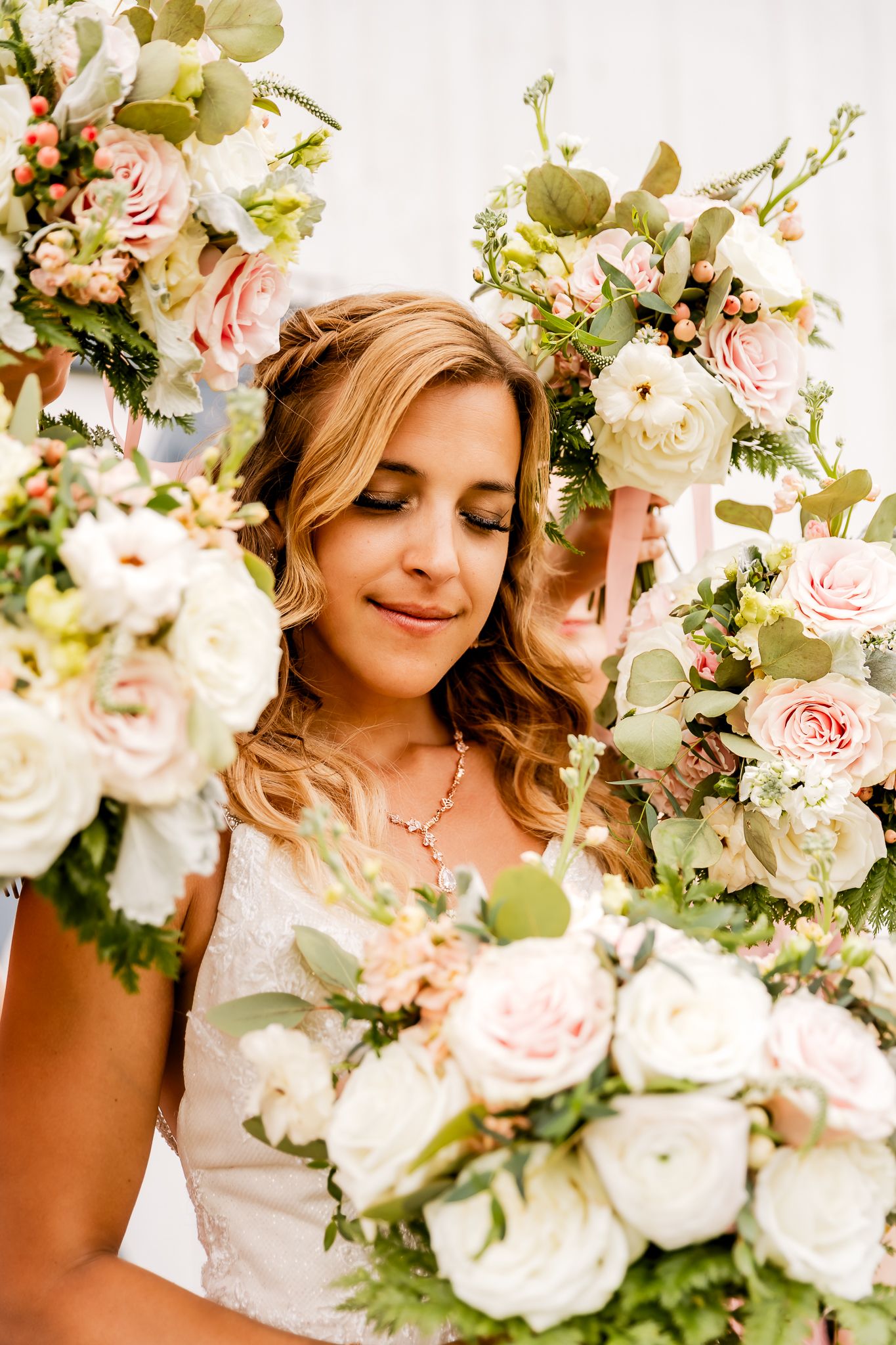 Bride surrounded by lush pink and white rose bouquets with eucalyptus greenery, eyes closed, wearing a white lace gown.