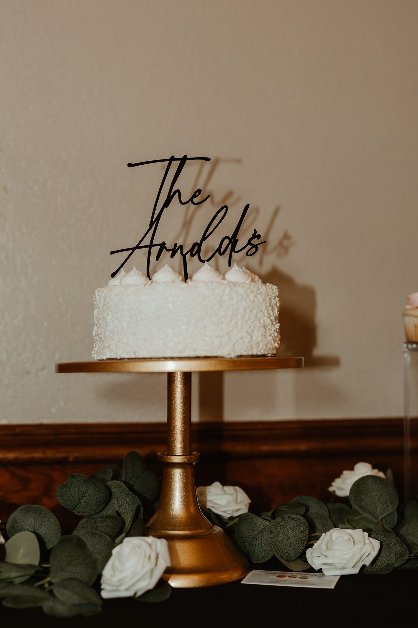 White wedding cake on gold stand with 'The Arnolds' topper, surrounded by greenery and white roses.