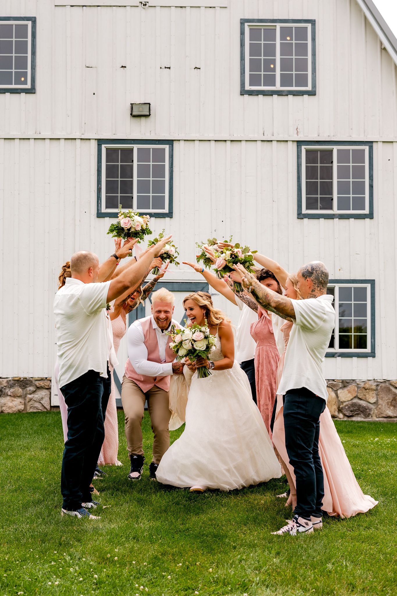 Wedding party celebrates outdoors, raising bouquets in arch over smiling bride and groom near white barn.