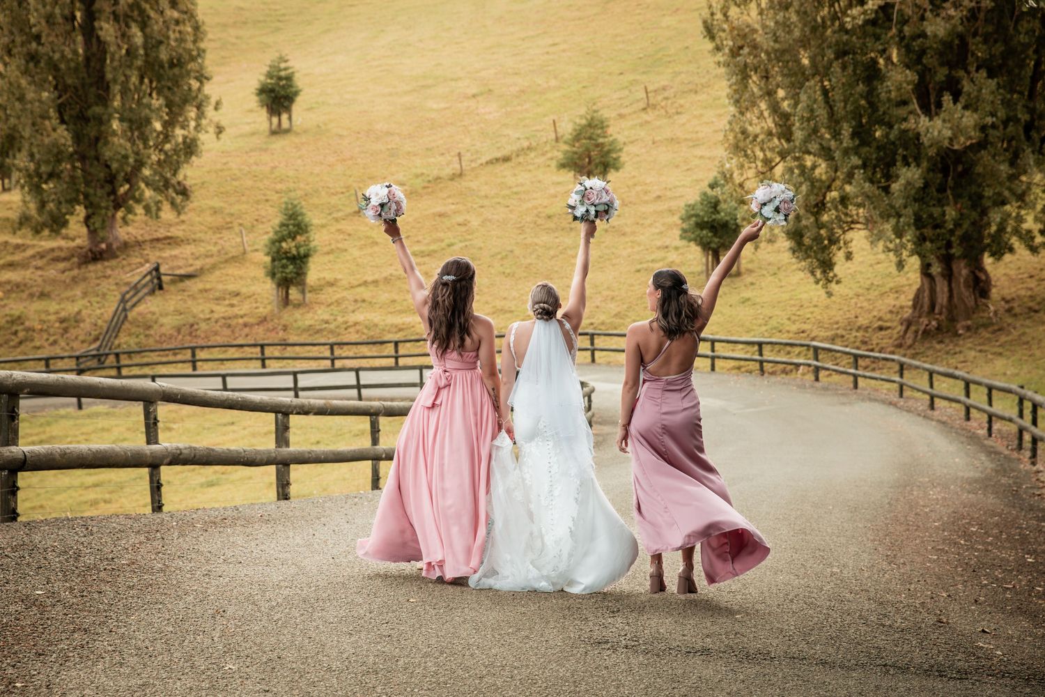 Bride and two bridesmaids in pink dresses raise bouquets walking along countryside path.