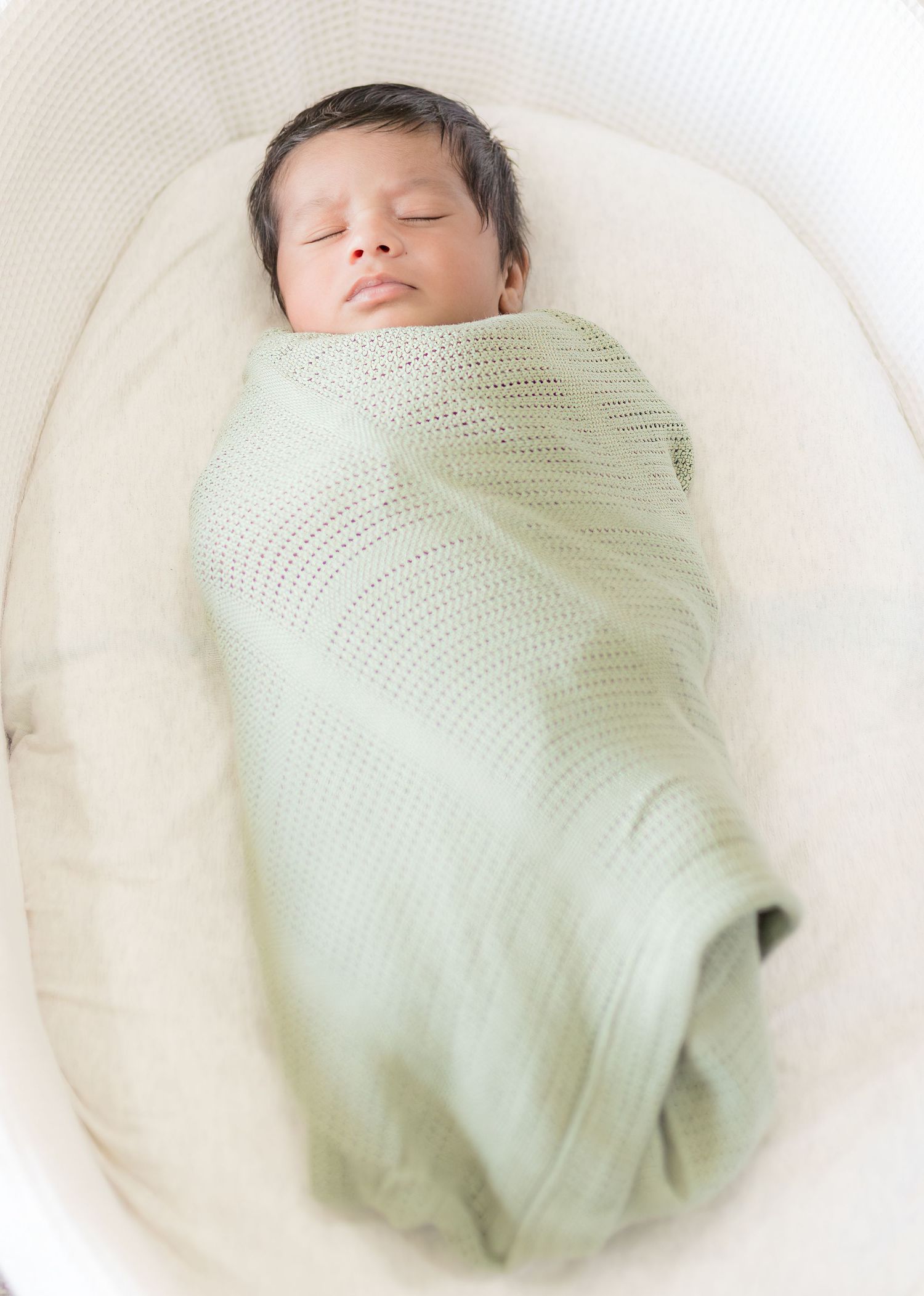 Sleeping newborn baby swaddled in a soft green knit blanket in a white bassinet.