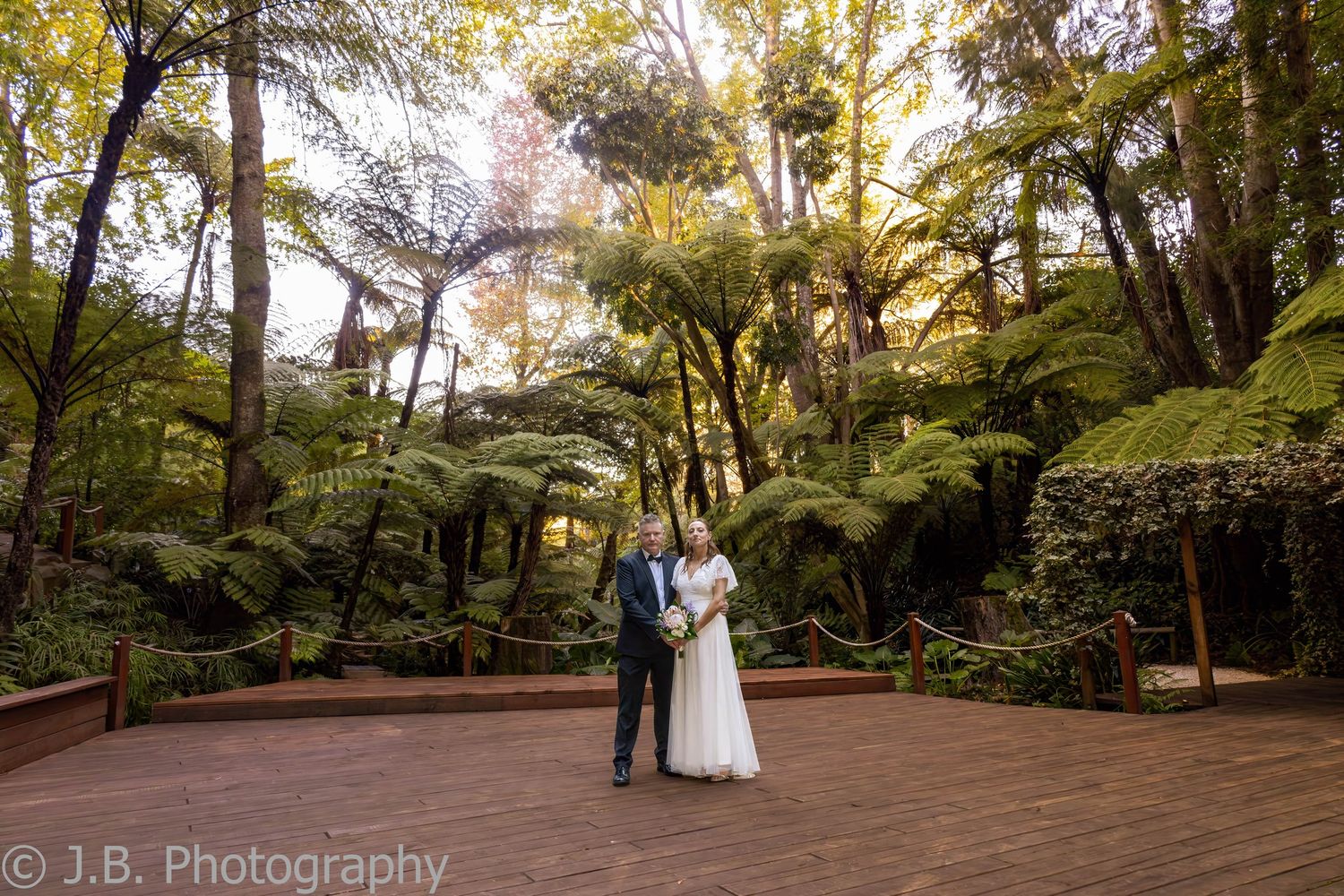 Wedding couple stands on wooden deck surrounded by lush fern tree forest in J.B. Photography shot.