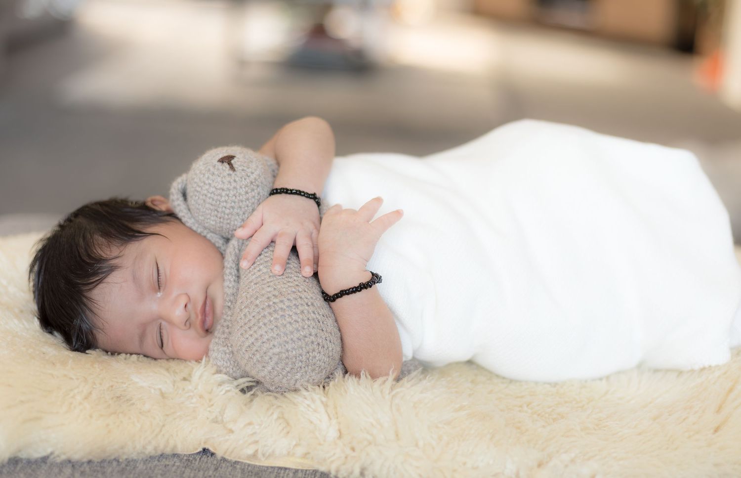 Peaceful newborn baby sleeping wrapped in white swaddle cuddling a knitted teddy bear on a fluffy cream rug.