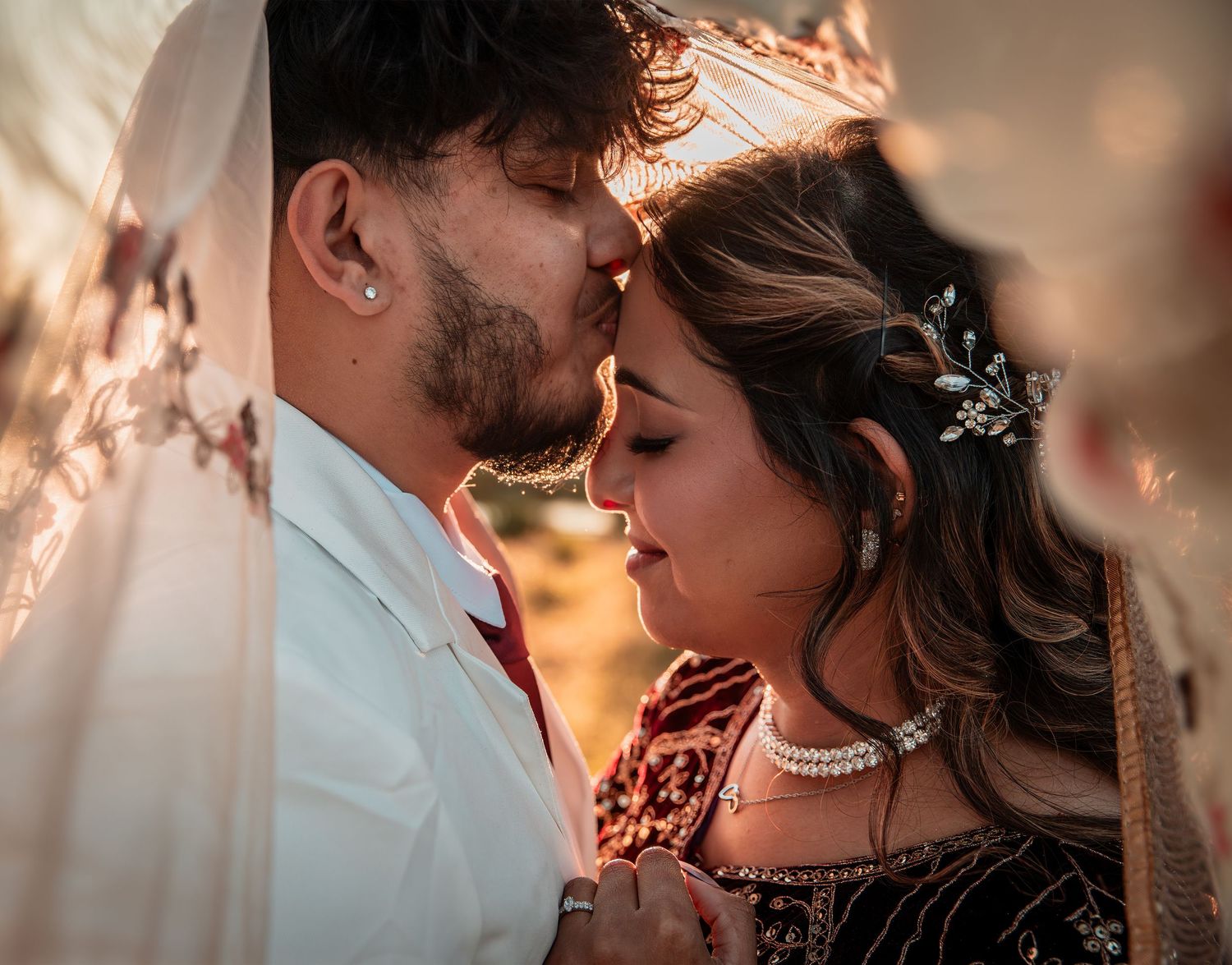 Indian couple in traditional wedding attire sharing an intimate moment, touching foreheads under a veil in golden light.