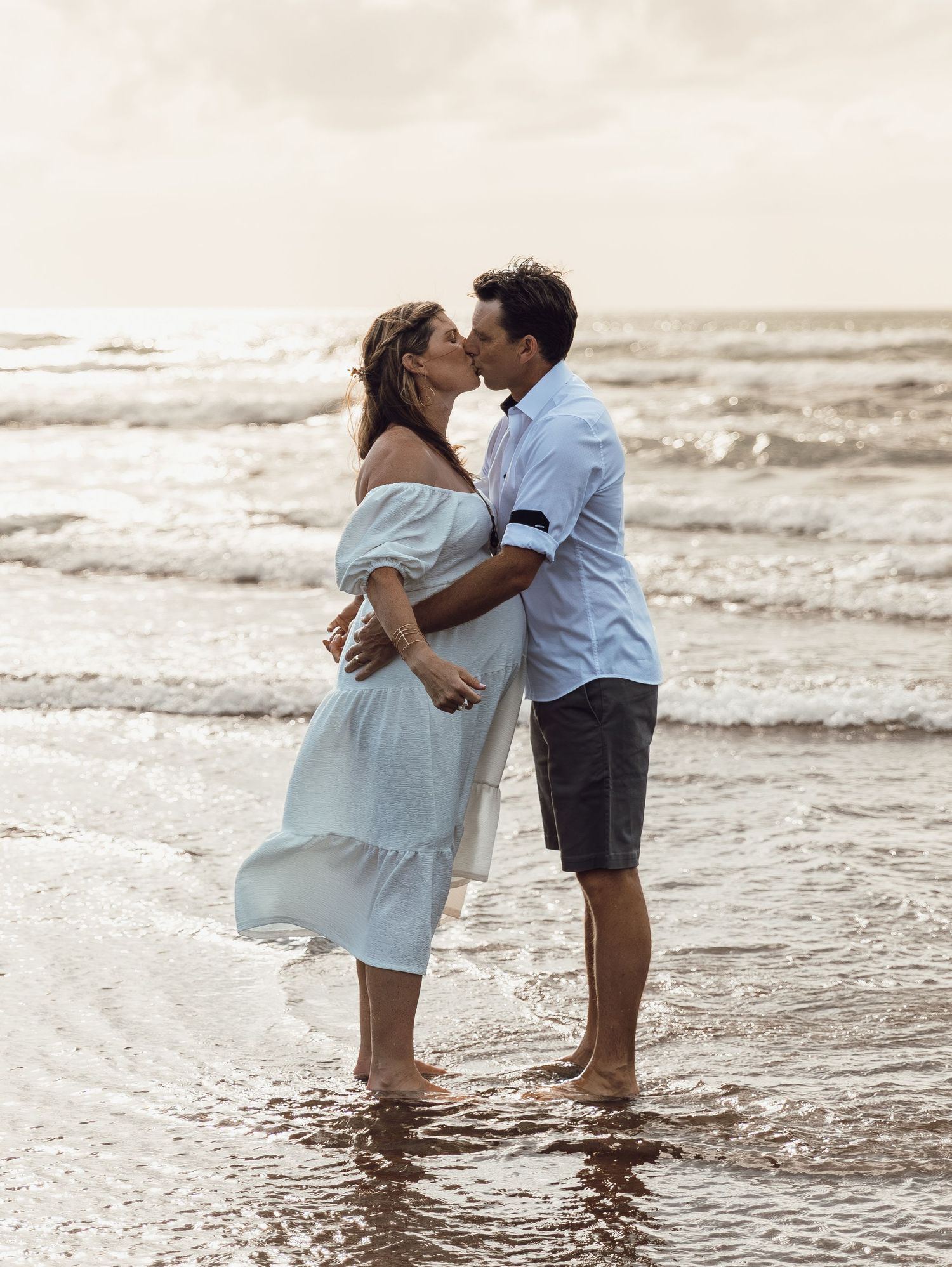 Couple sharing a romantic kiss while standing barefoot in ocean waves on a beach during golden hour.