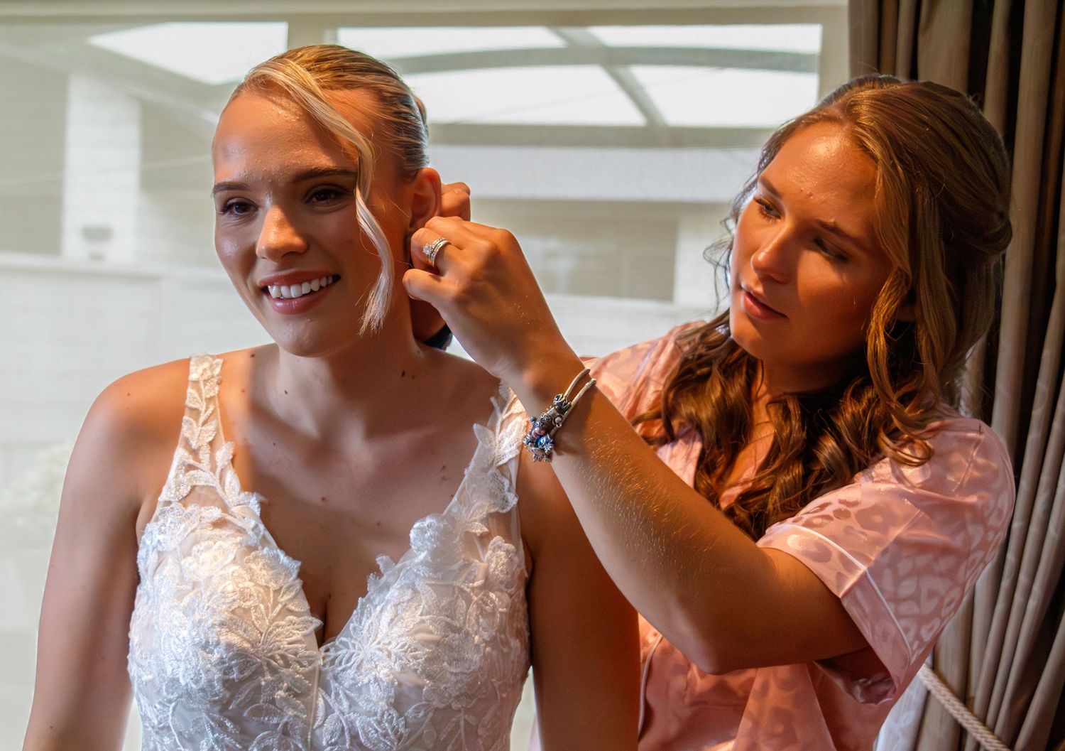 Bridesmaid helping smiling bride put on earrings while getting ready on wedding morning.