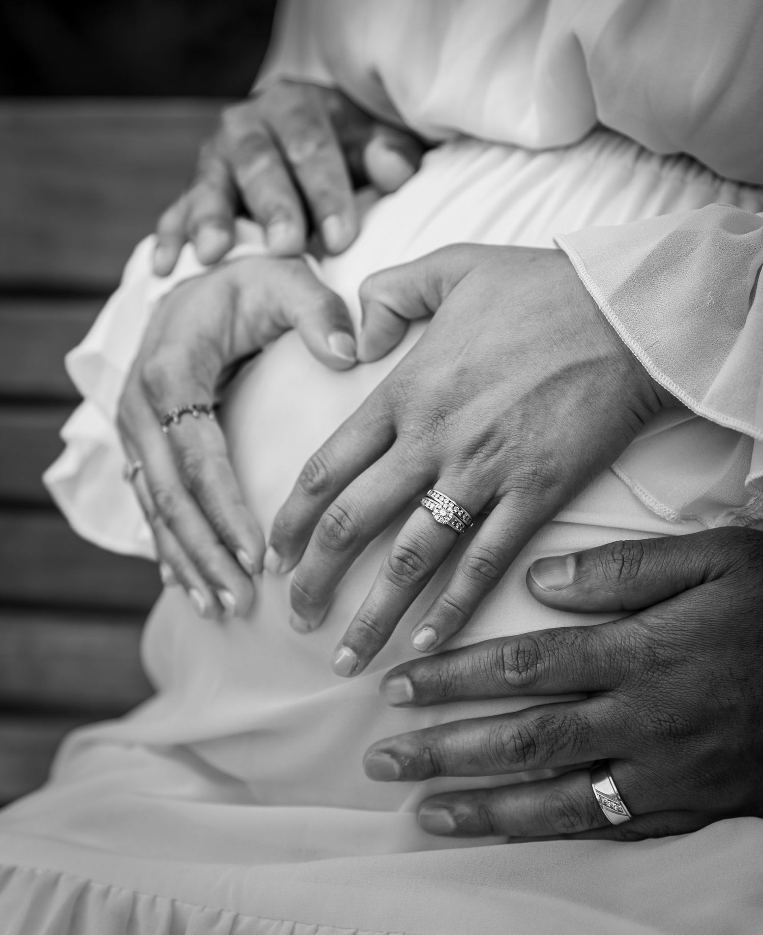 Black and white close-up of couple forming heart shape with hands on pregnant belly, wearing wedding rings.