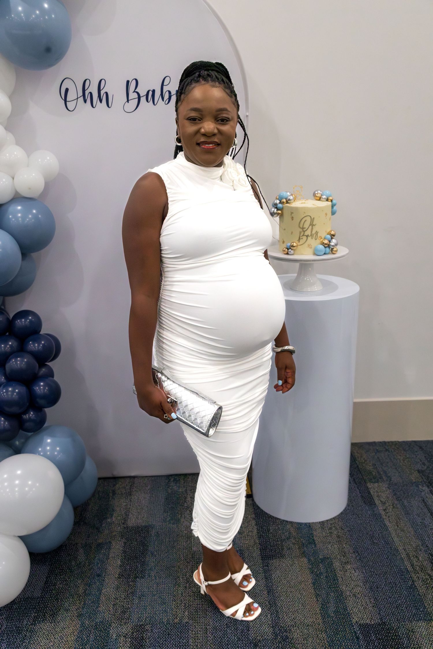 Pregnant woman in white bodycon dress posing at baby shower with blue balloon decorations and cake.