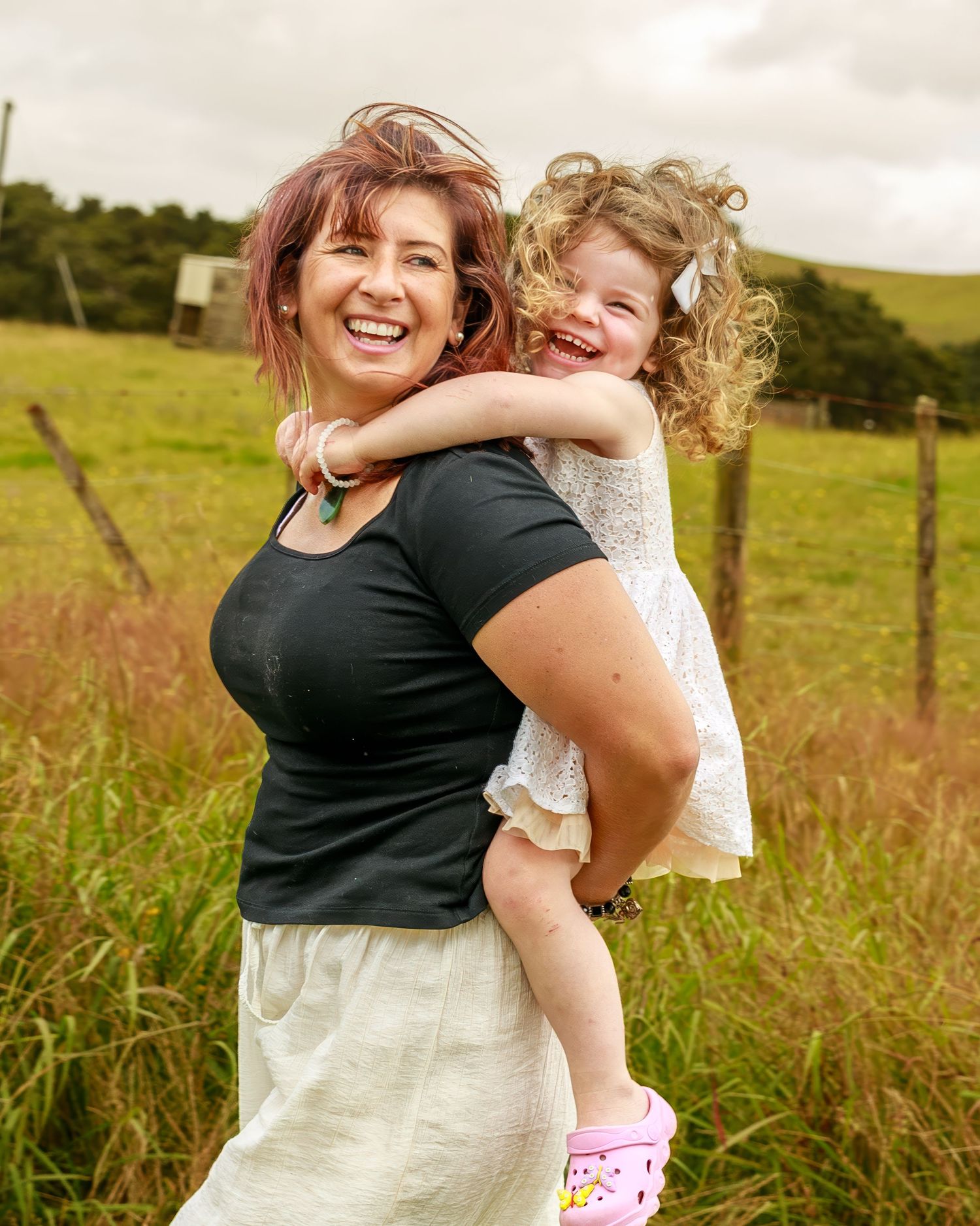 Joyful mother carrying laughing curly-haired daughter on back in outdoor field portrait.