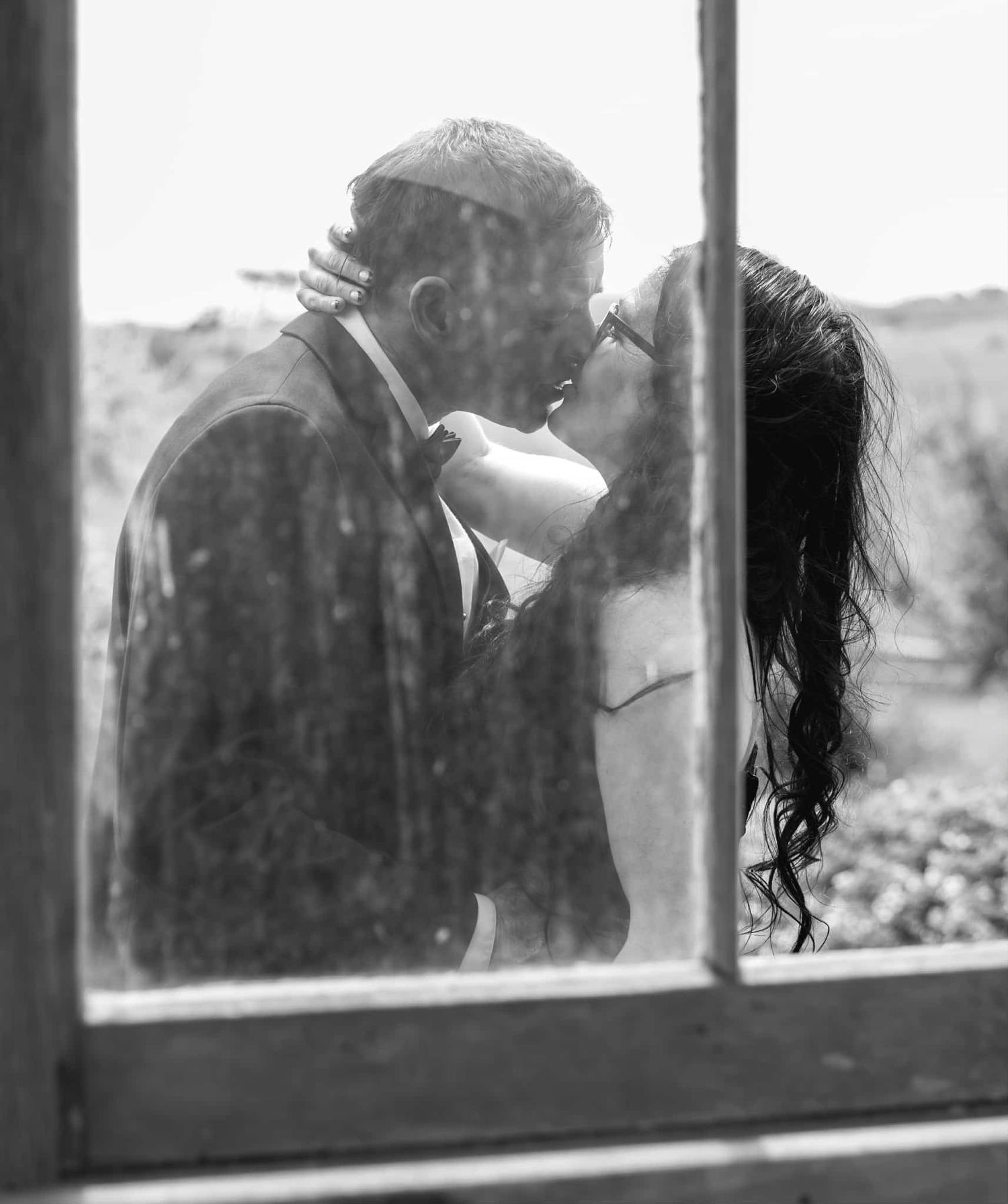 Romantic black and white photo of a couple kissing viewed through a rustic window pane.