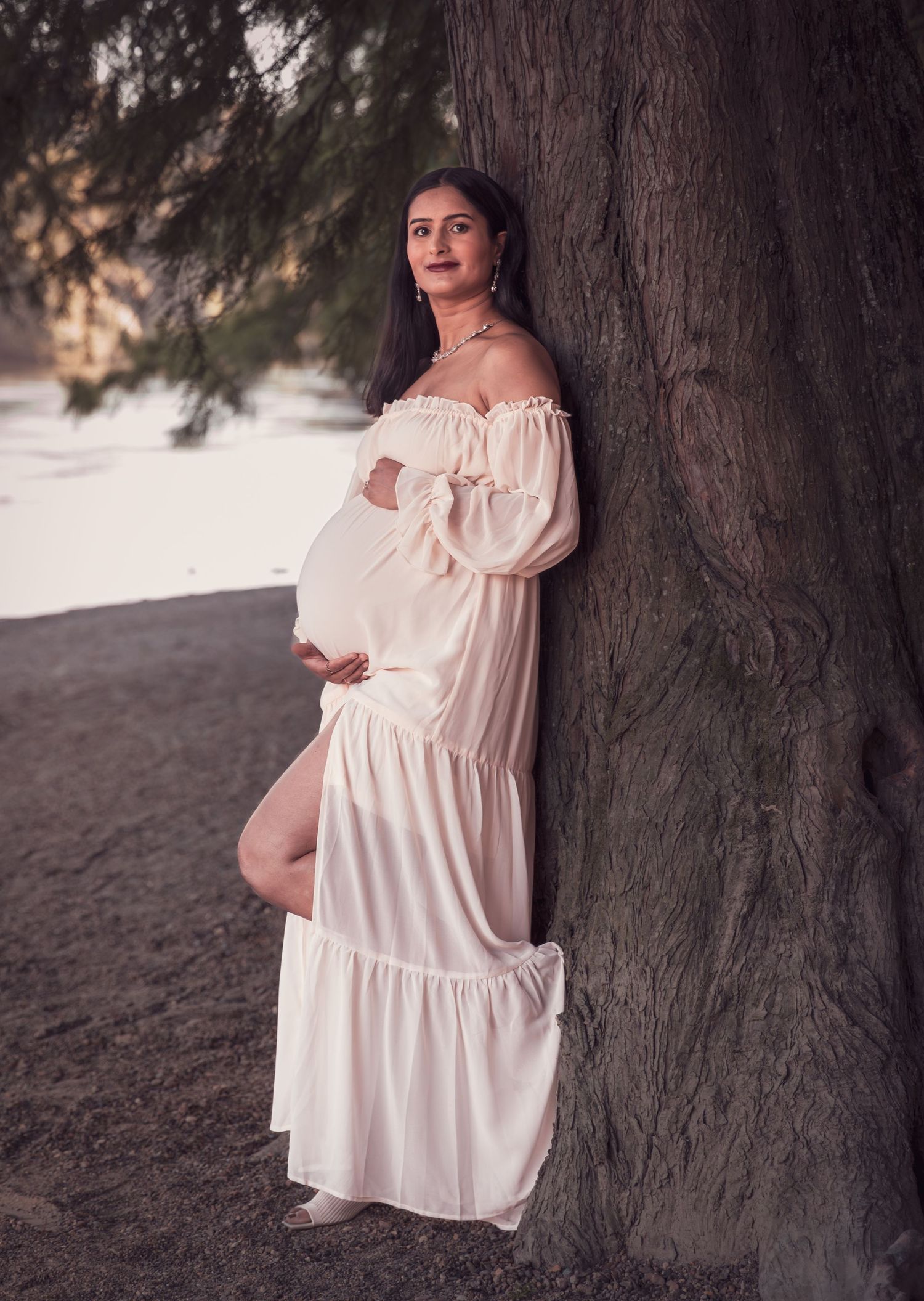 Pregnant woman in flowing white off-shoulder dress leaning against a large tree in a serene outdoor setting.