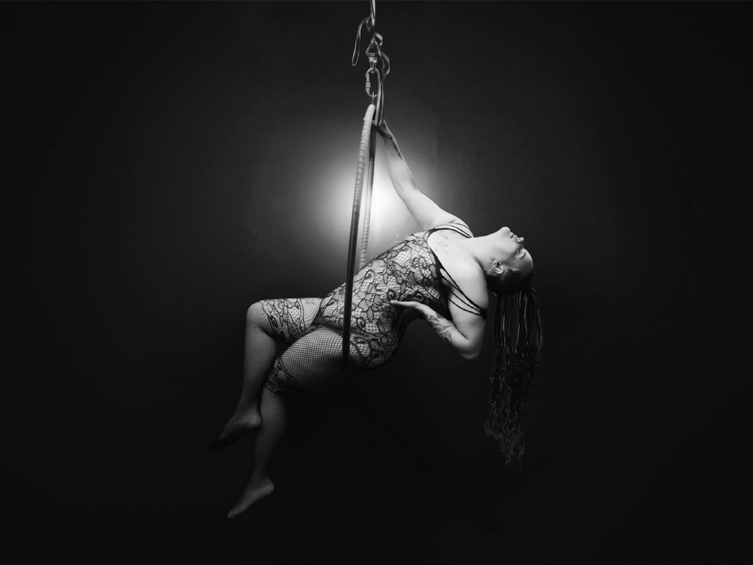 Black and white artistic photo of a tattooed woman performing an aerial pole dance in dramatic lighting.