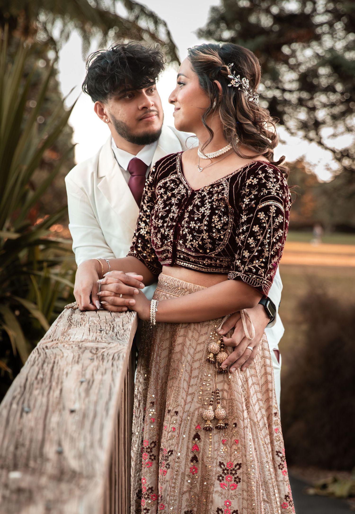 Indian couple in traditional wedding outfits posing outdoors, woman in embroidered lehenga and man in white suit.