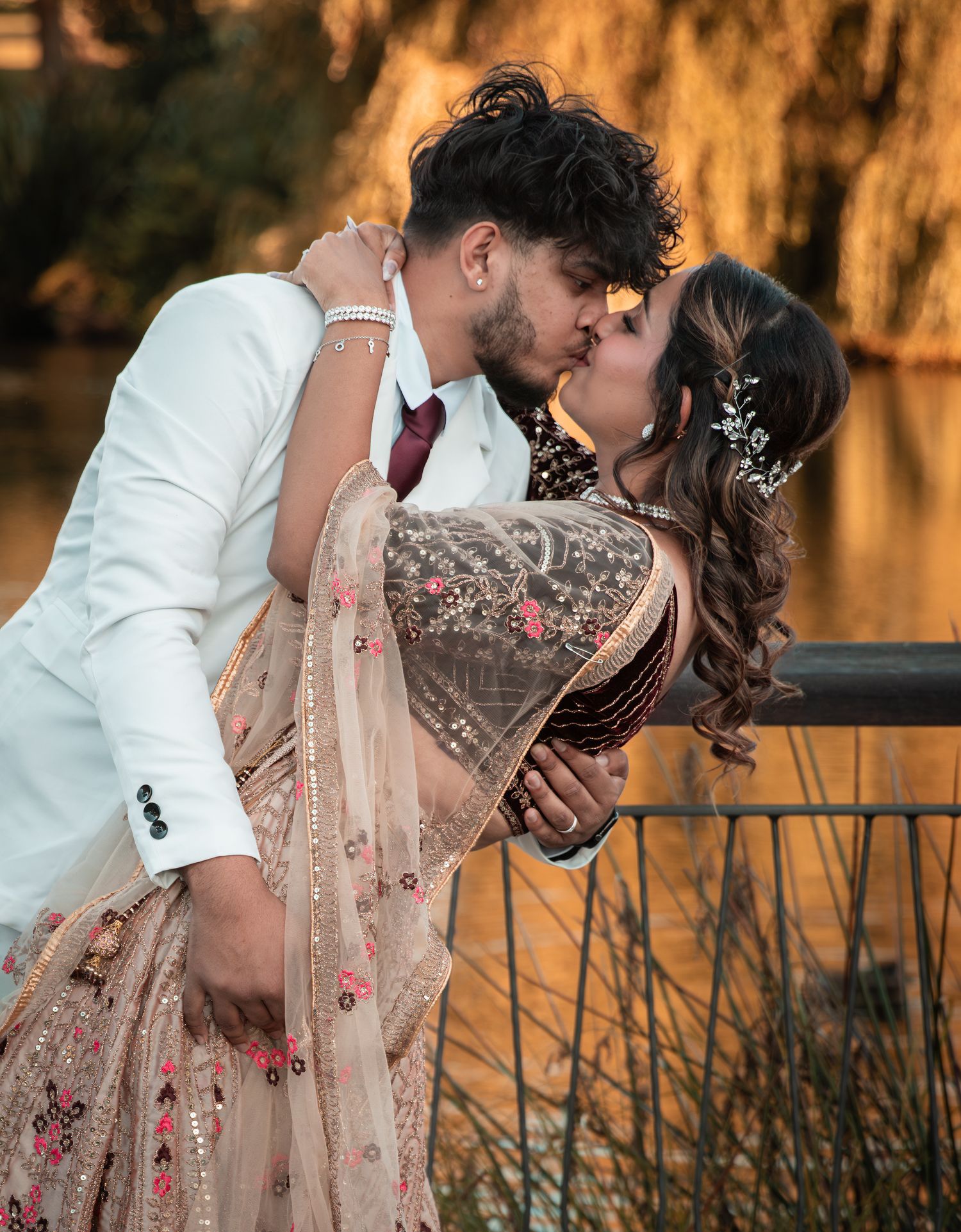 Couple sharing a romantic kiss at sunset wearing traditional Indian wedding attire near a lake.