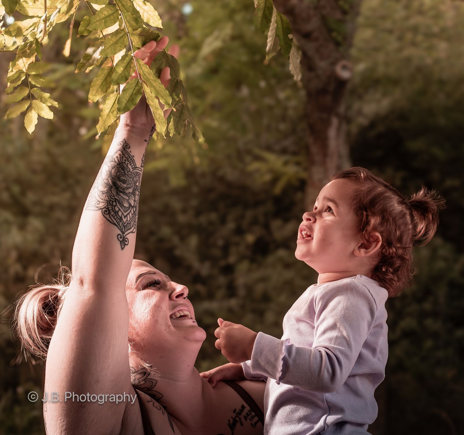 Mother with arm tattoo lifts laughing toddler toward tree branches in golden light.