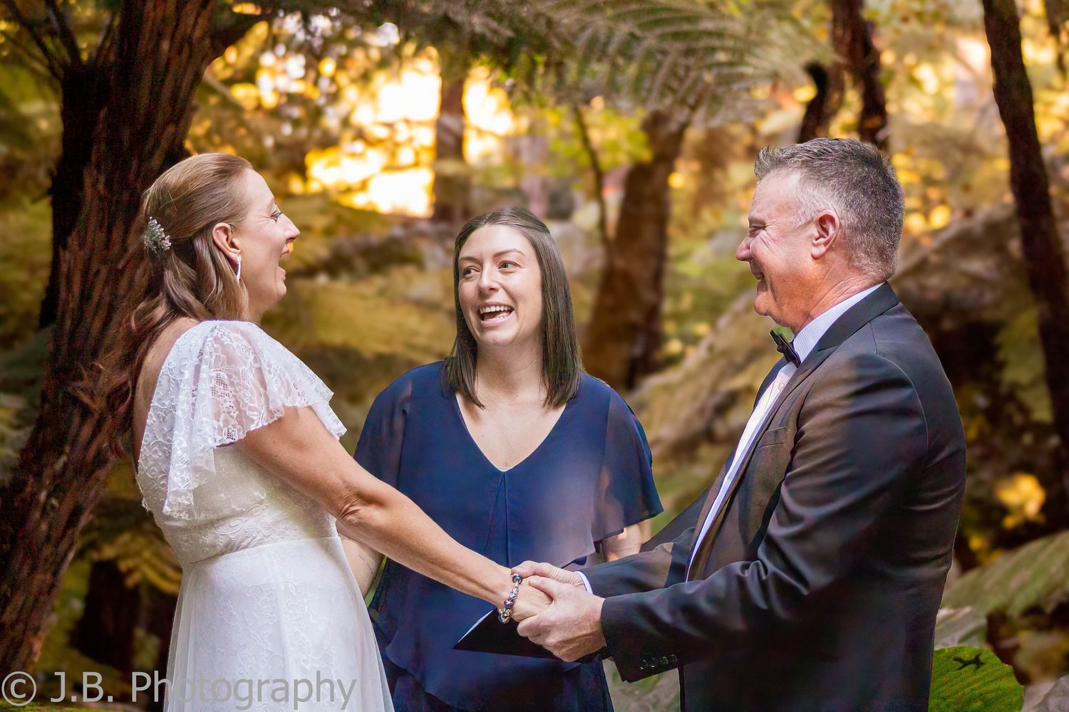 Bride and groom hold hands laughing with celebrant during outdoor forest wedding ceremony.