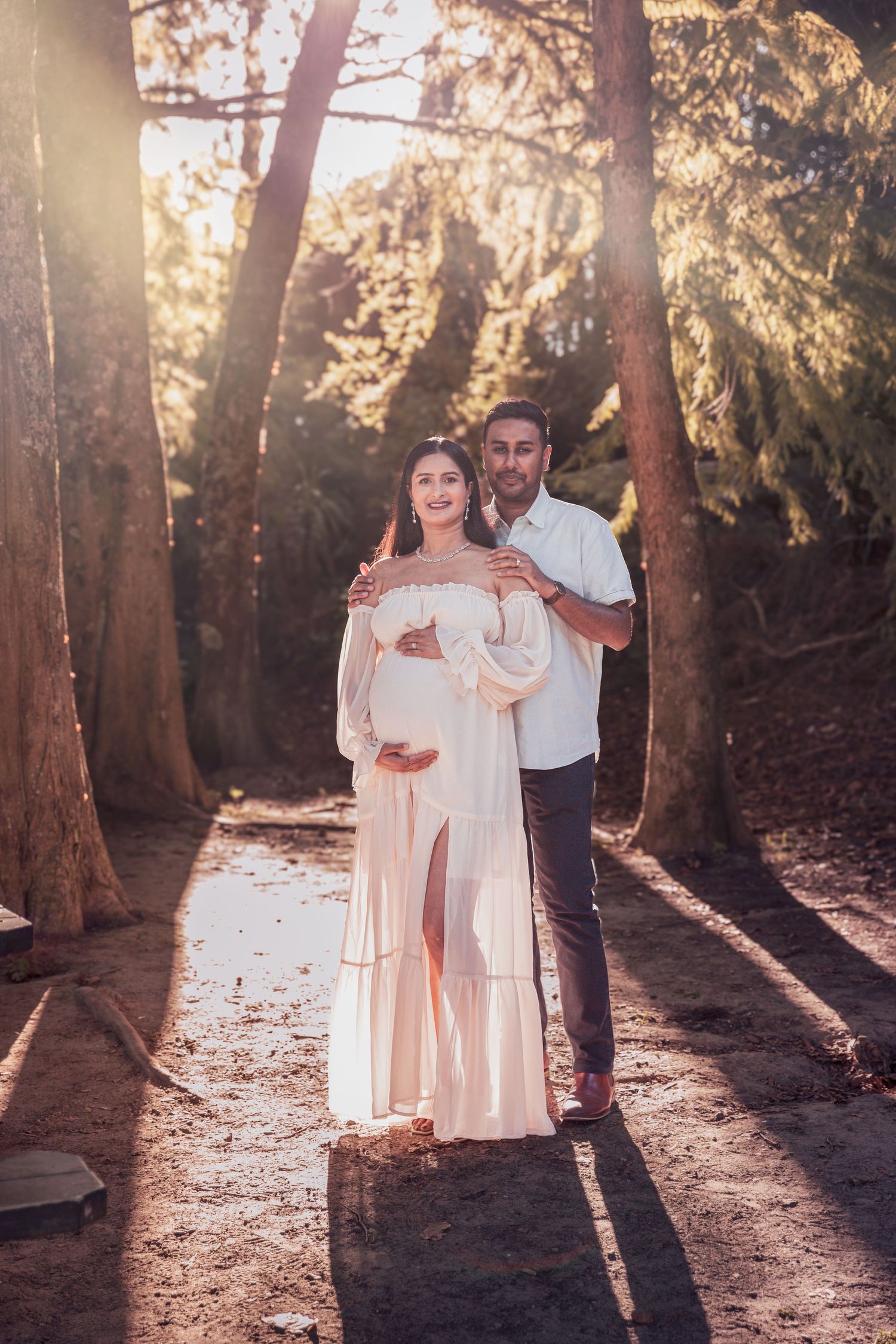 Pregnant couple poses in golden sunlight among tall trees, woman in flowing white maternity gown.