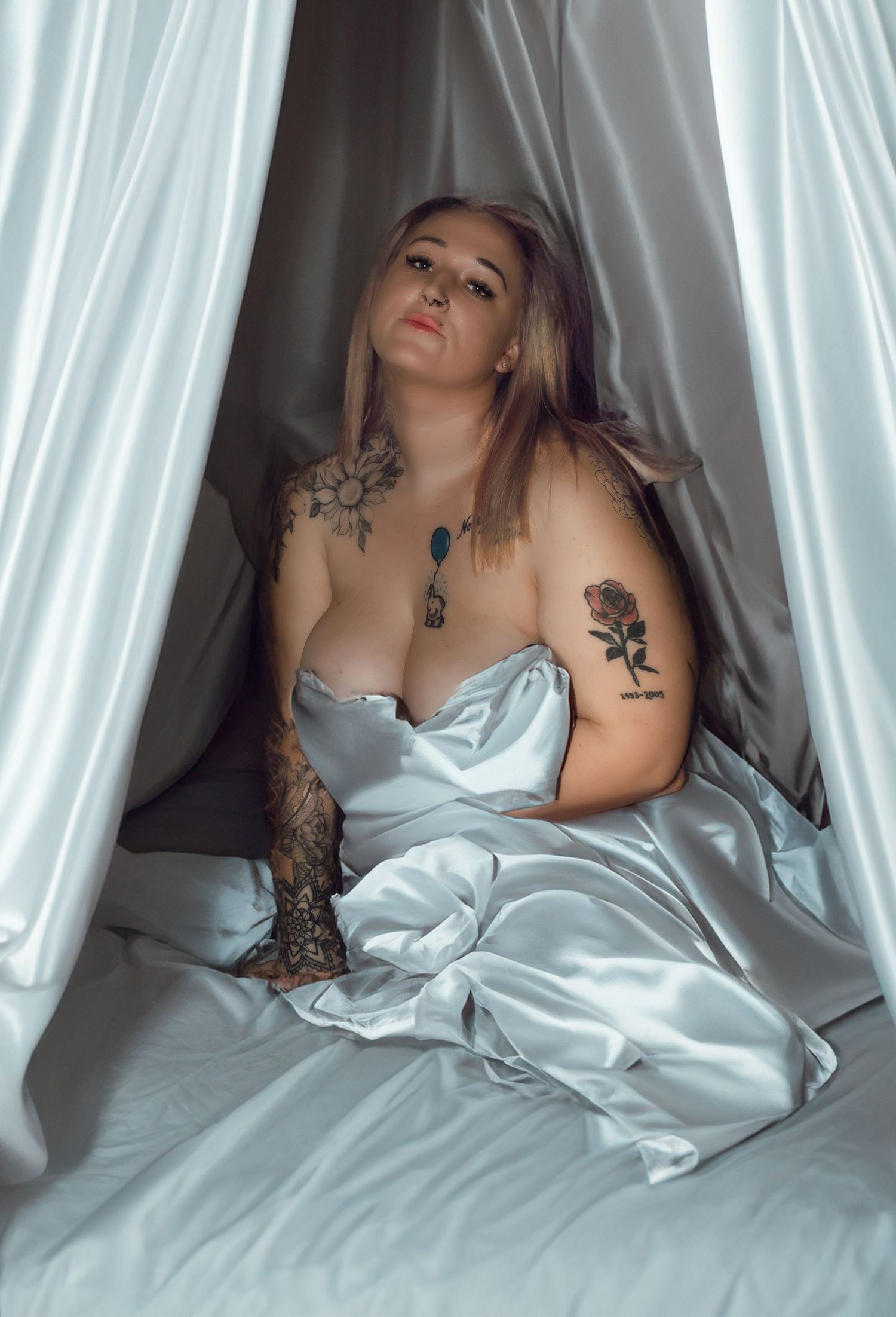 Tattooed woman wrapped in white satin sheets in artistic boudoir photography portrait.