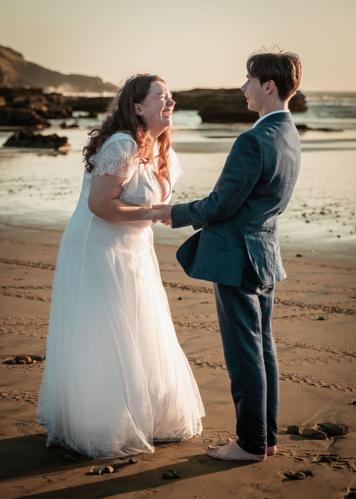 Bride and groom holding hands and laughing together on a sandy beach at golden hour sunset.