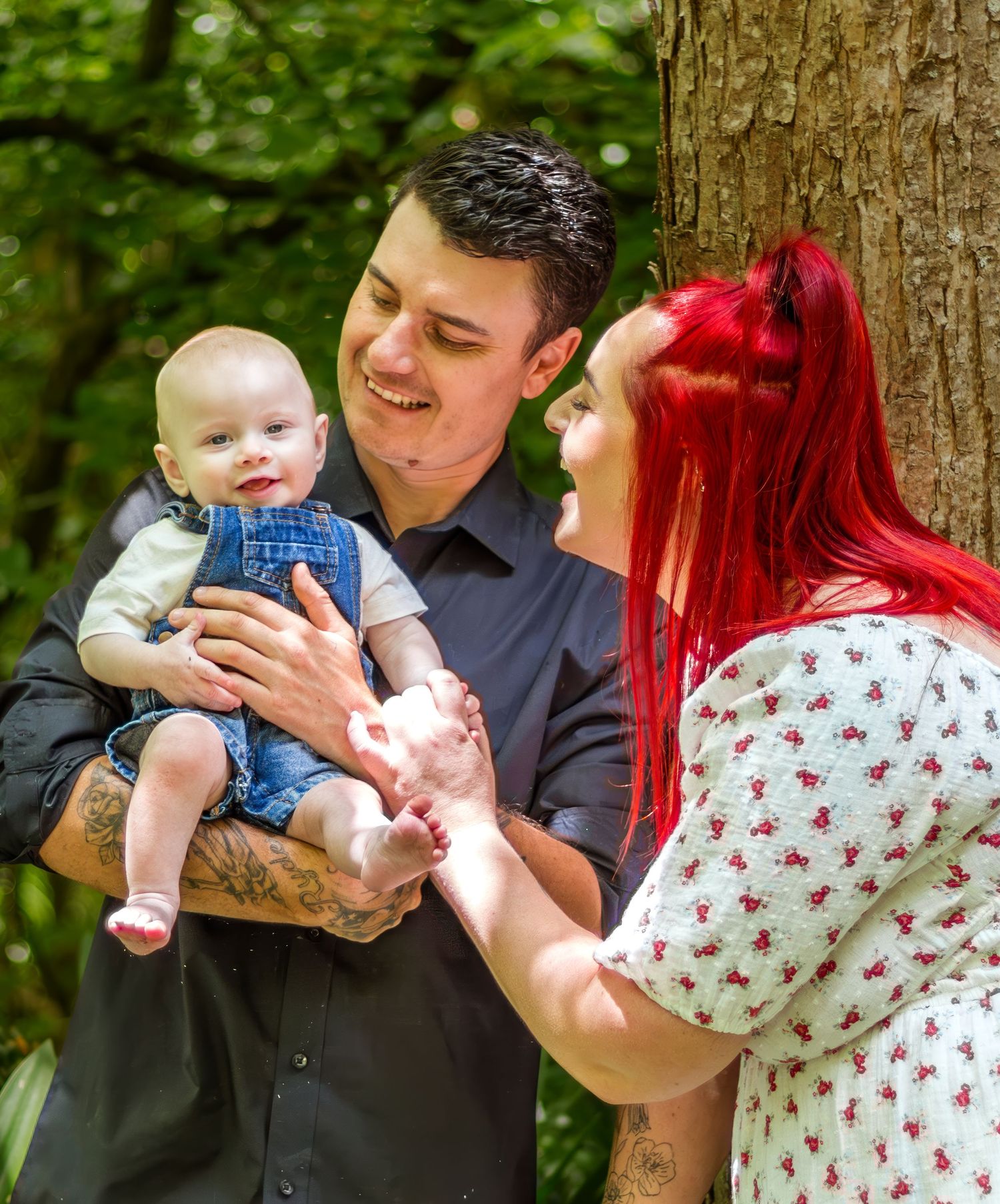 A couple with a tattooed man and red-haired woman lovingly interact with their baby outdoors.