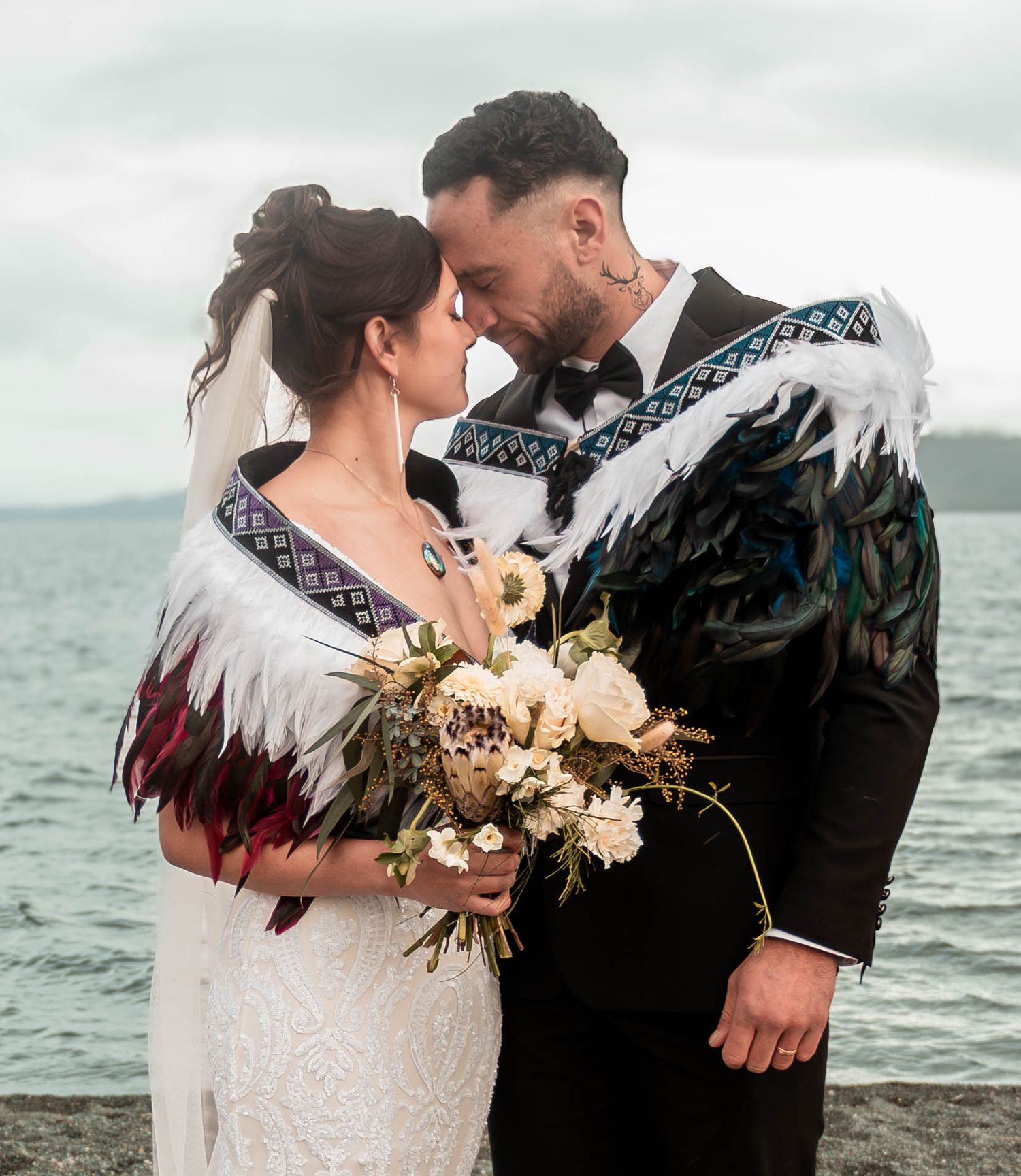 Couple in traditional Maori feather cloaks share intimate moment by waterfront on their wedding day.