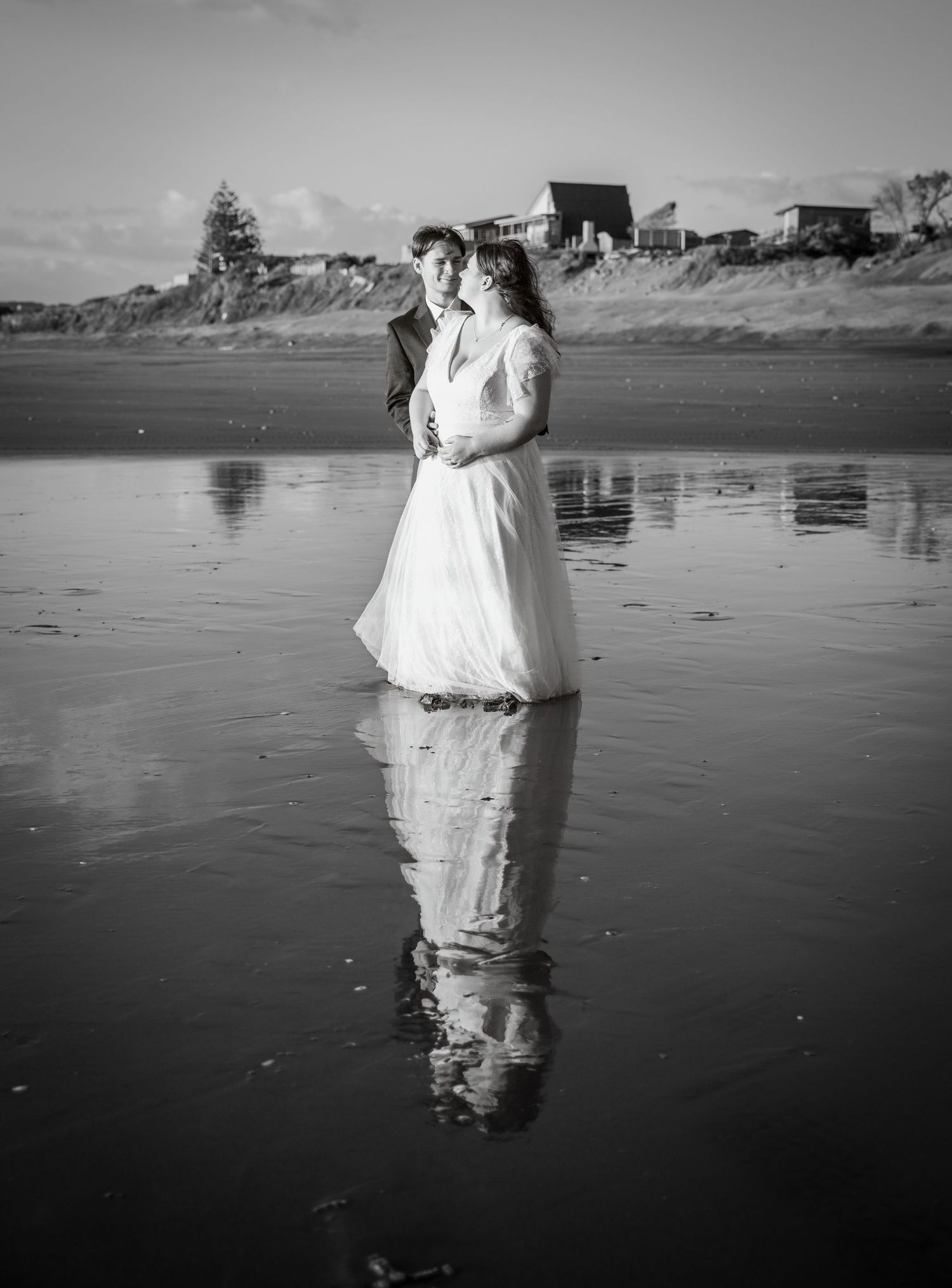 Bride and groom standing on reflective wet beach, stunning black and white wedding portrait.