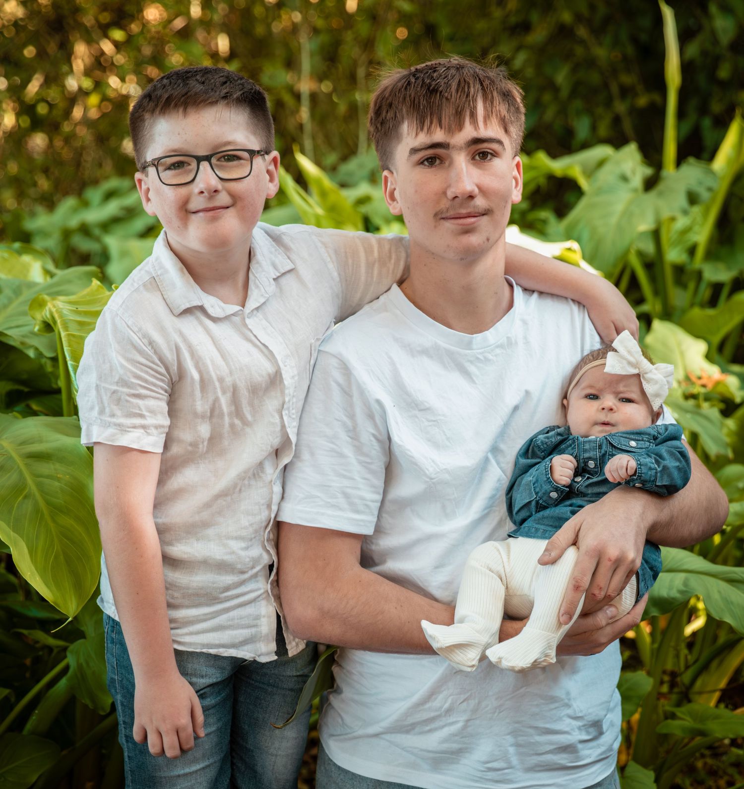 Young man holding newborn baby with older brother posing in lush garden family portrait.