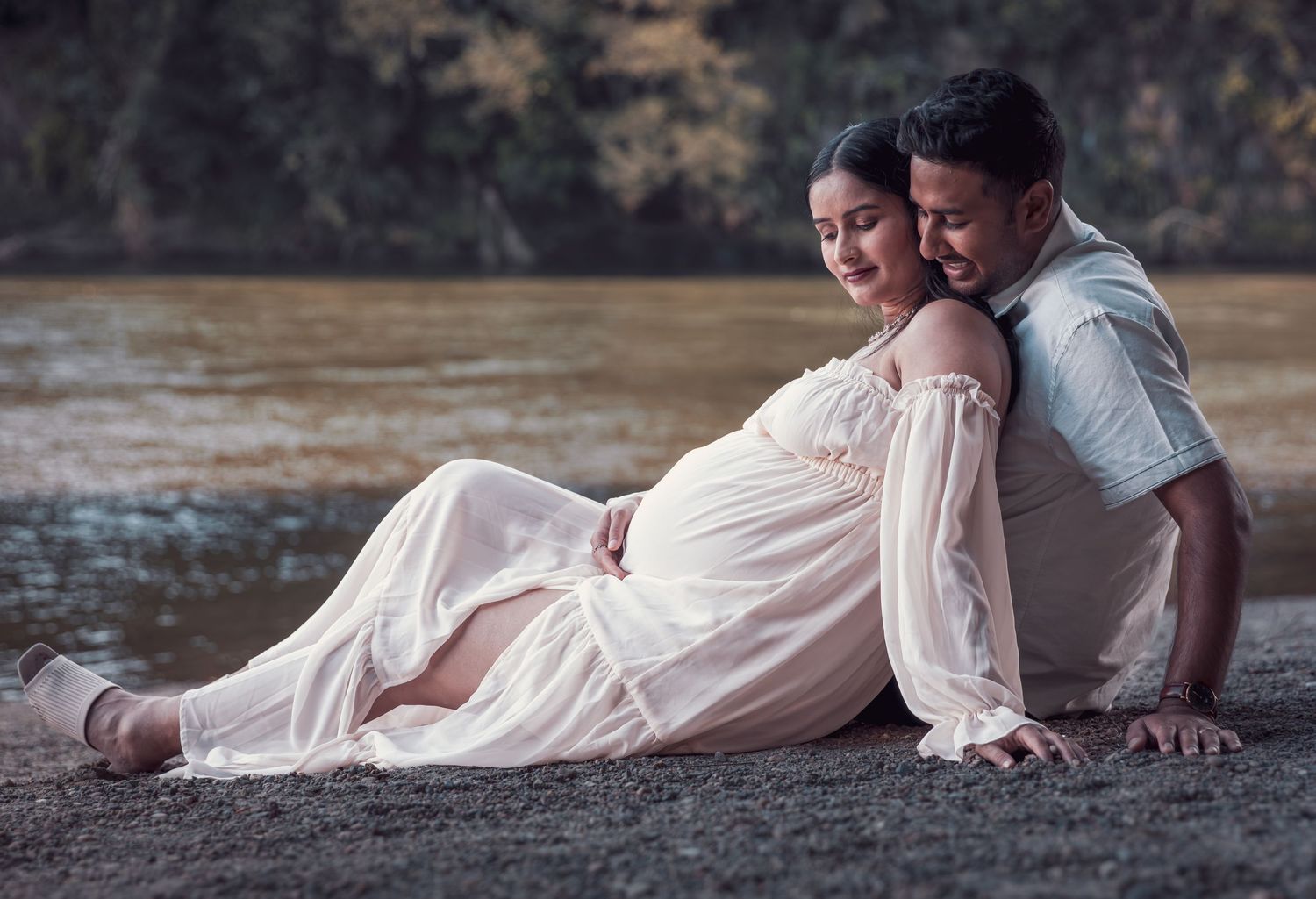 Romantic maternity photo of couple sitting by riverside, woman in white dress leaning against partner.