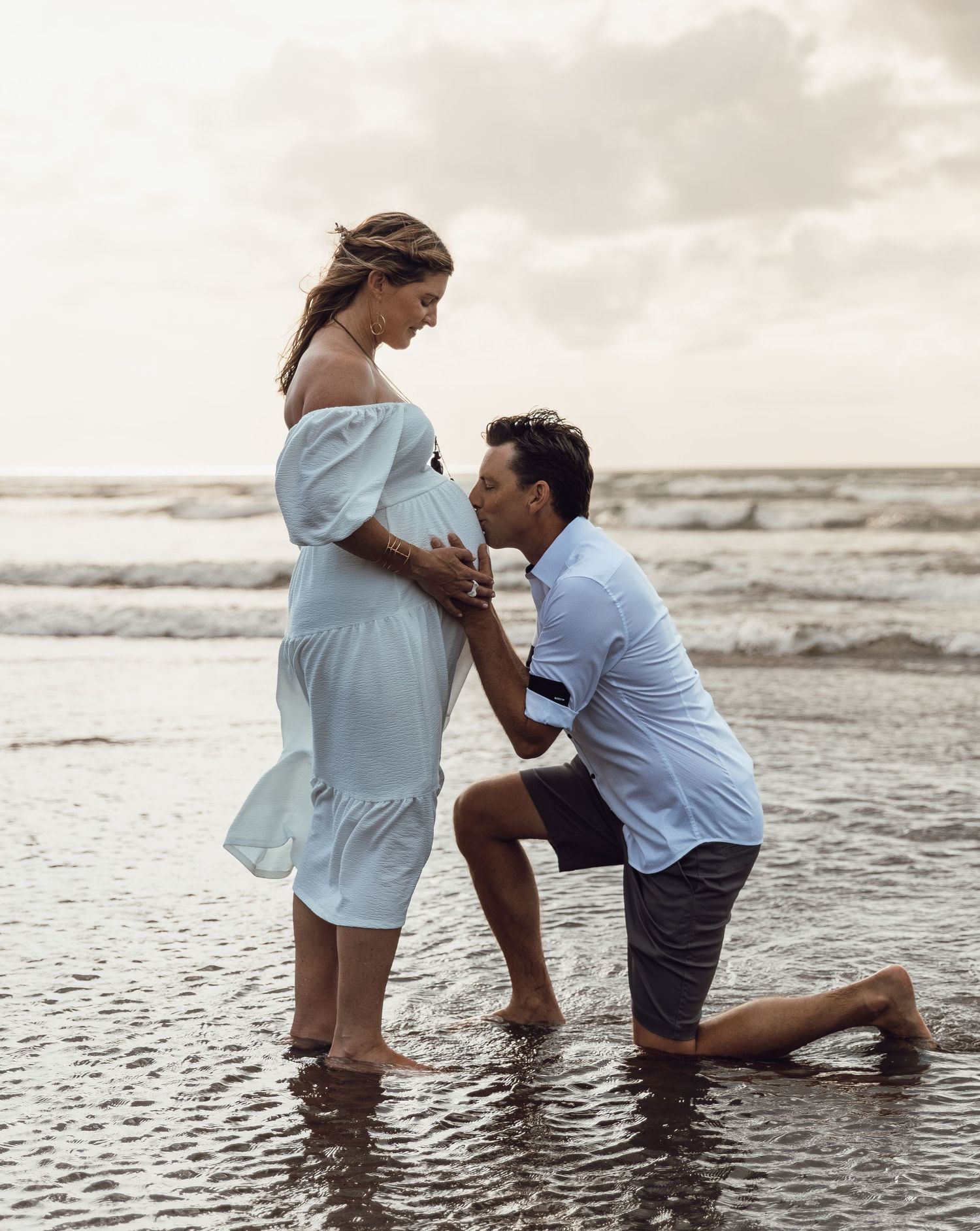 Man kneeling in shallow ocean water kissing pregnant woman's baby bump during a maternity beach photoshoot.