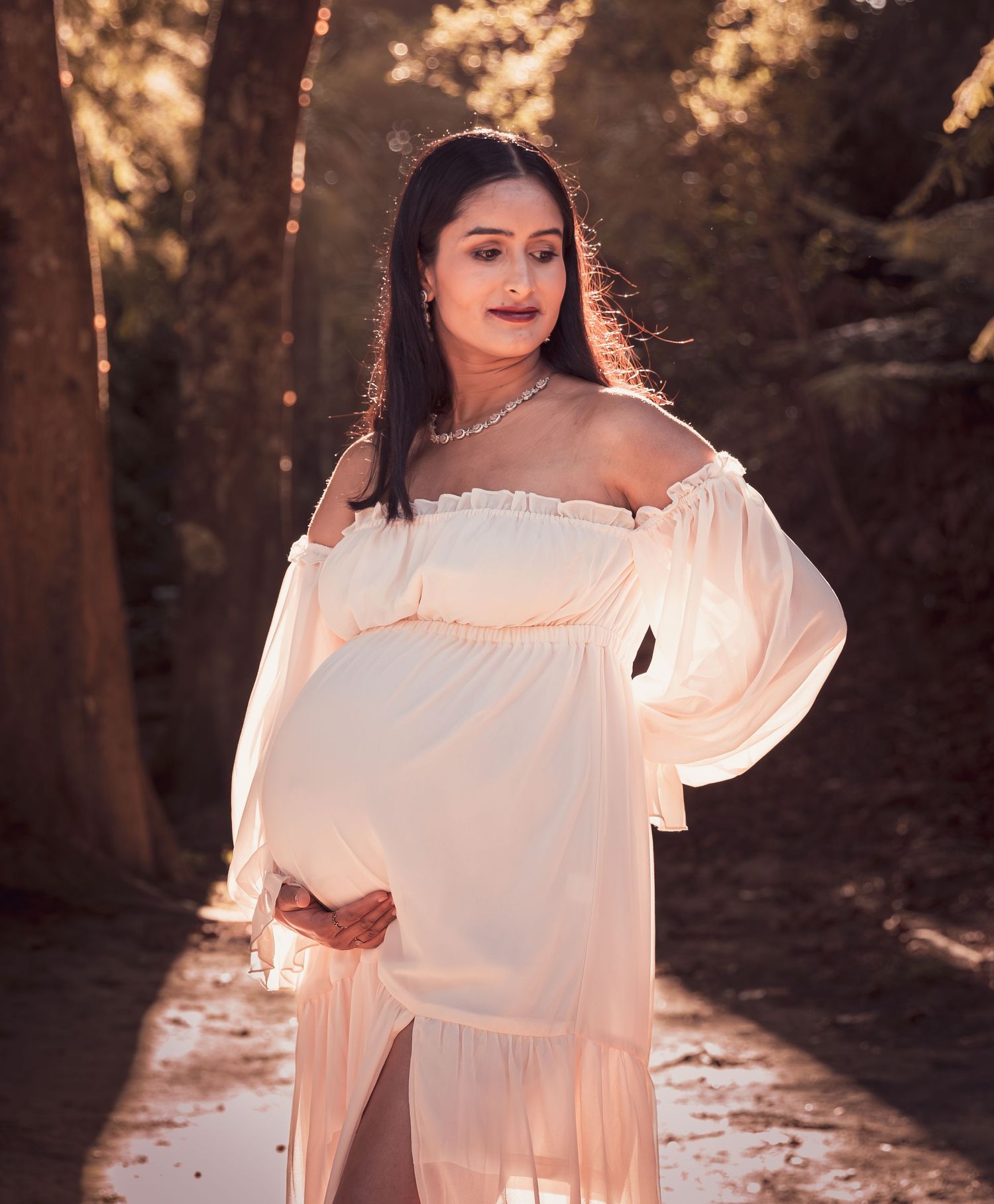 Pregnant woman in elegant white off-shoulder maternity gown posing outdoors in golden sunlight.