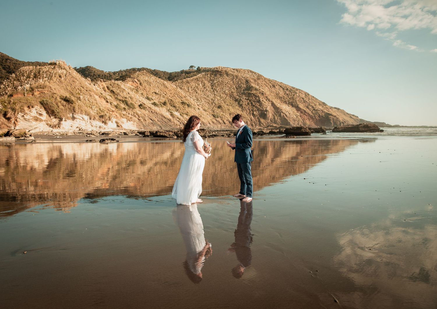 Bride and groom standing on a reflective wet beach with golden cliffs in the background during their wedding photoshoot.