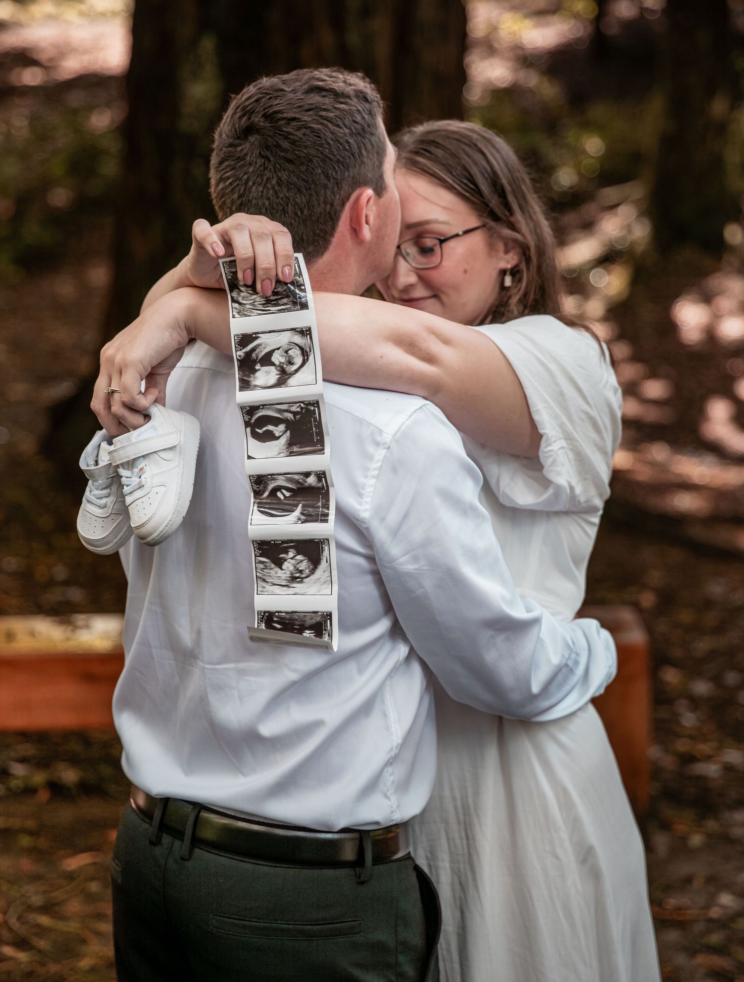 Couple embracing during pregnancy announcement, woman holding ultrasound strip and tiny baby shoes.