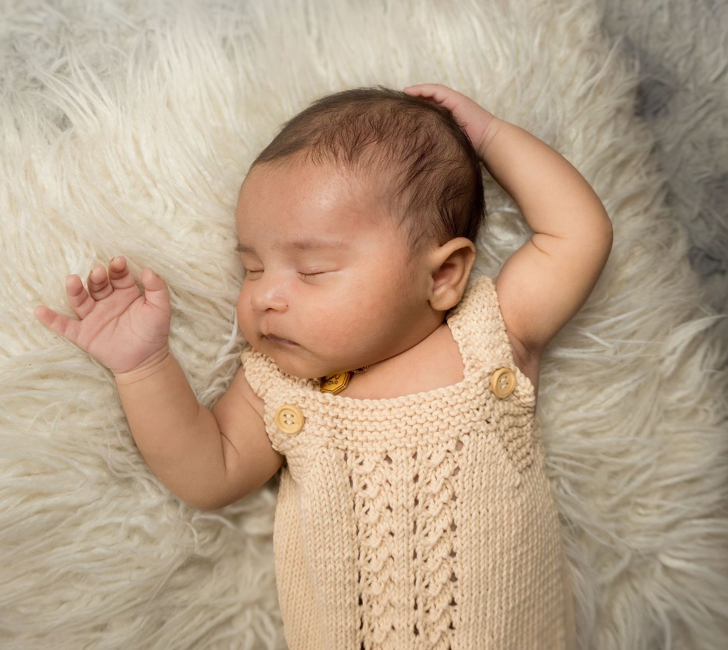 Sleeping newborn baby in cream knitted overalls resting on a fluffy white fur rug with arm raised.