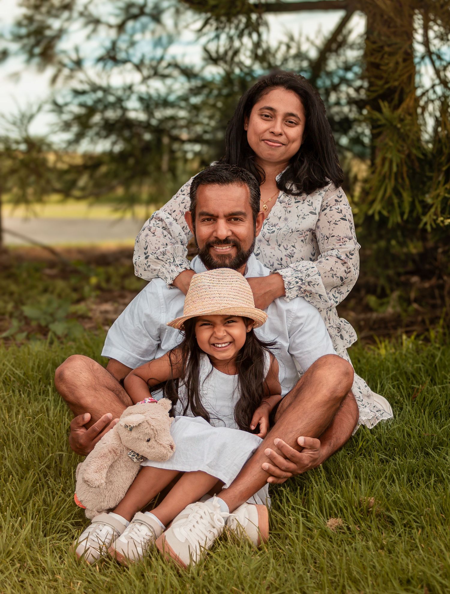 A happy family of three sits together on green grass with a young girl in front.