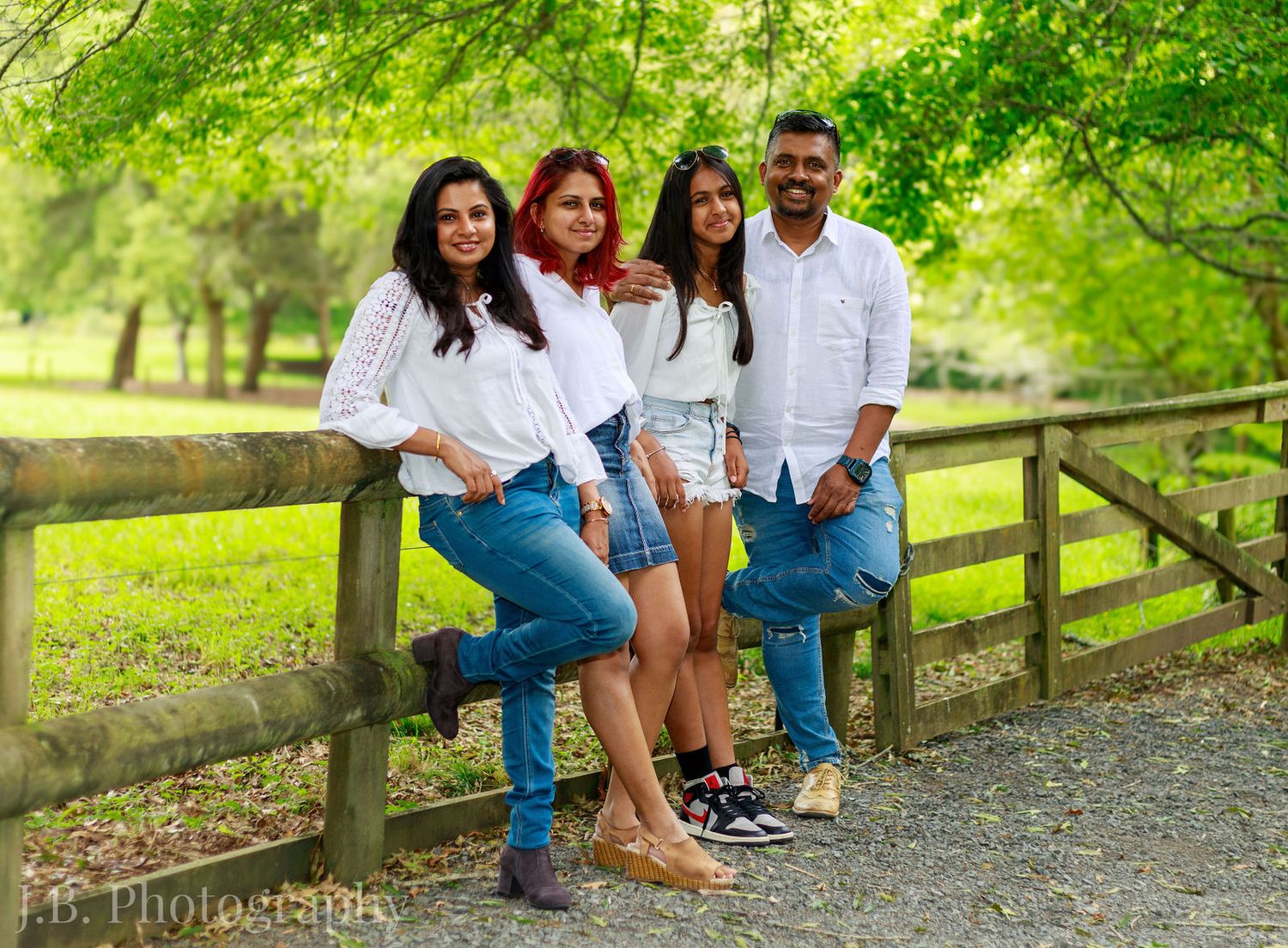 Family of four poses by wooden fence in park, wearing white tops and jeans for outdoor portrait session.