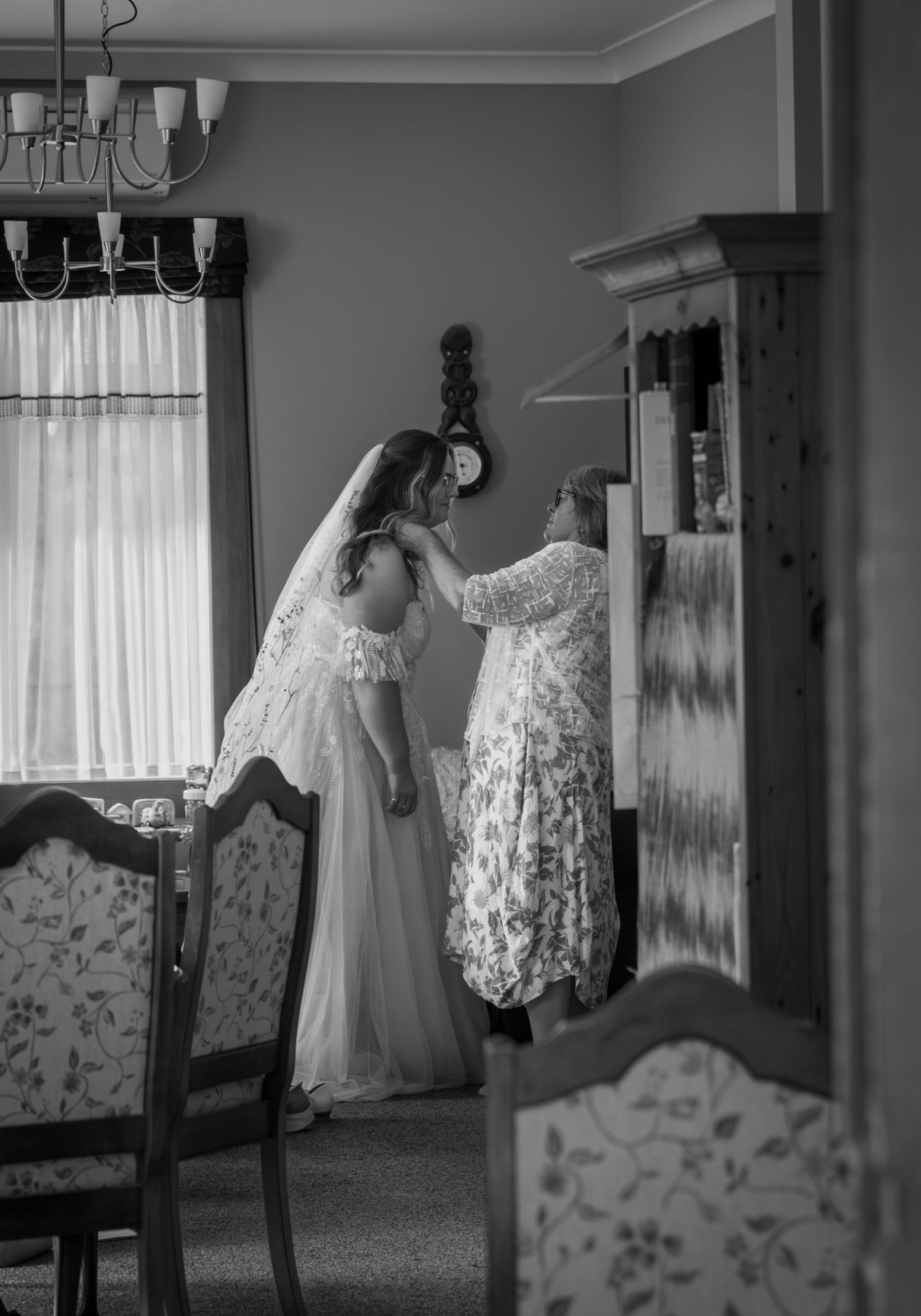 Mother helping bride adjust wedding veil in vintage dining room, black and white photo.