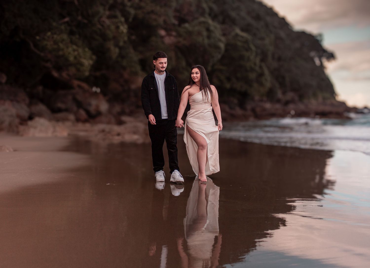 Couple walking hand in hand along reflective beach shore at sunset in elegant attire.