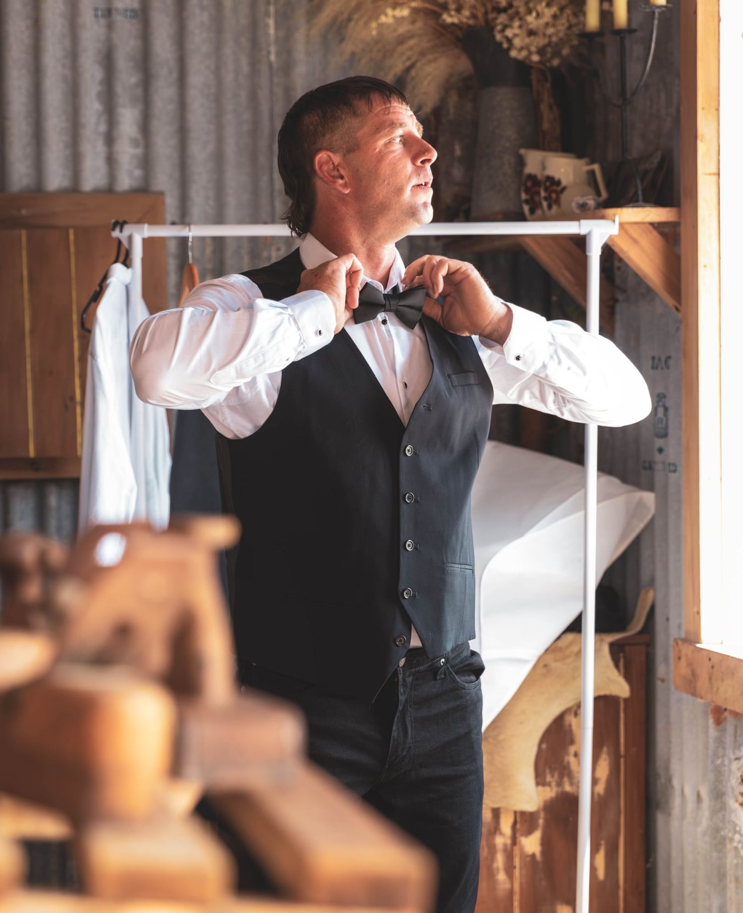 Groom adjusting bow tie while getting ready in a rustic barn setting before his wedding.
