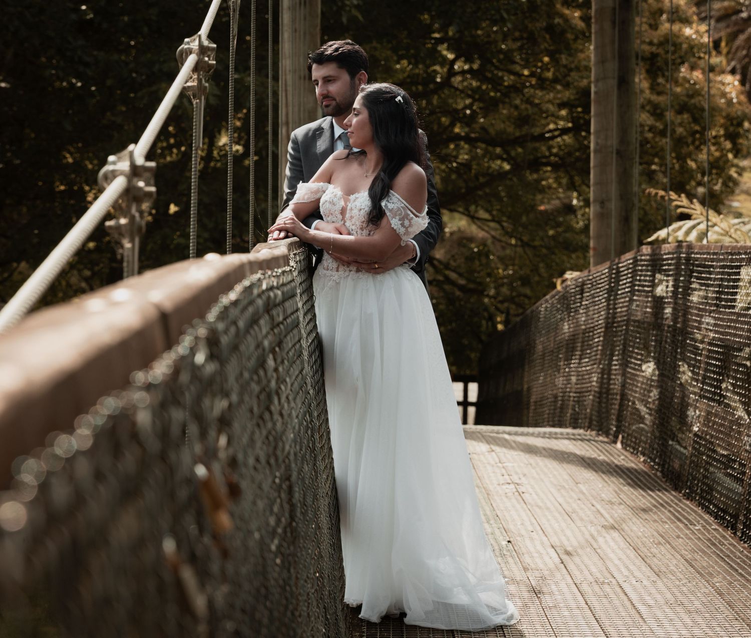 A bride and groom embrace on a wooden suspension bridge surrounded by lush greenery on their wedding day.